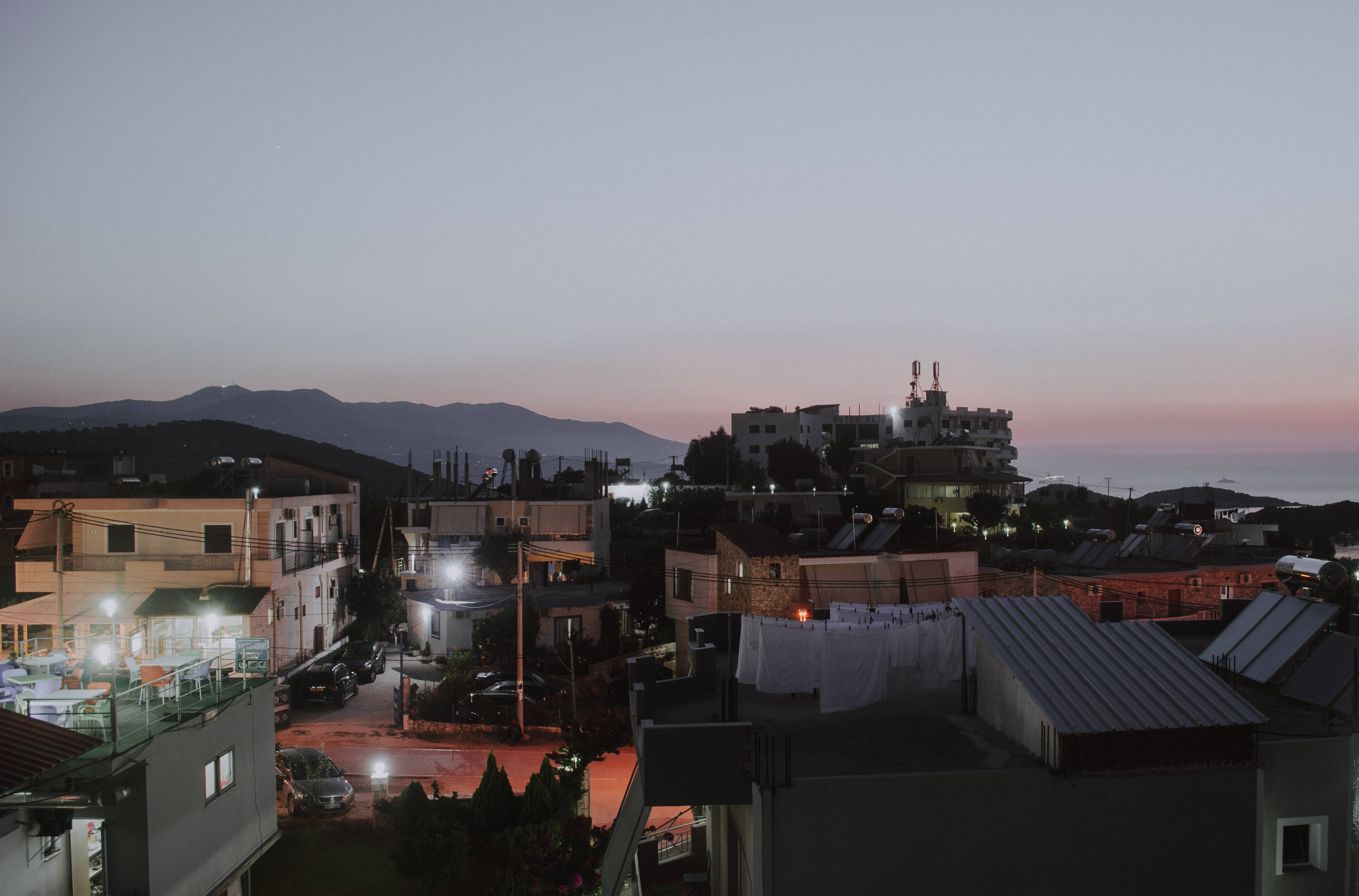 Albania 桌布 壁紙 — 暮色中的沿海小鎮，溫暖的路燈在遠處山巒上方柔和的粉紅色和藍色天空中閃耀。