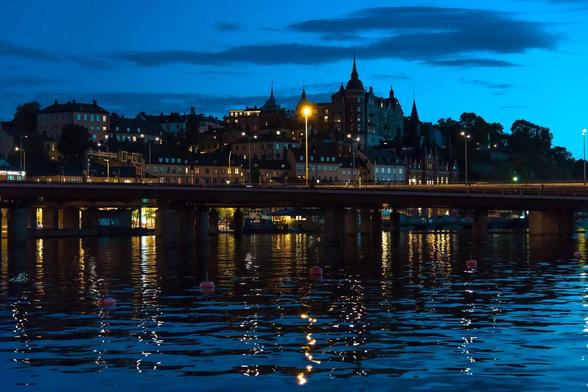 Sweden 桌布 壁紙 — 暮色城市景觀，一座橋樑橫跨水面，倒映著歷史建築在深藍色天空下的溫暖燈光。