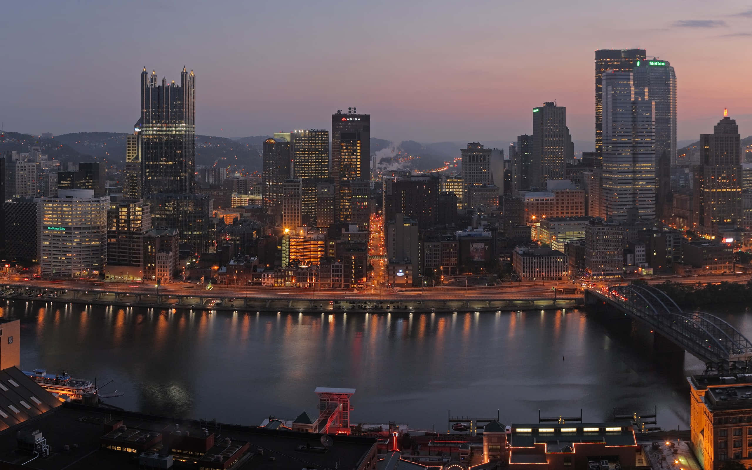 Pittsburgh Skyline 桌布 壁紙 — 暮色城市景觀，在柔和粉紅與紫色天空下，發光的摩天大樓倒映在平靜的河面上。