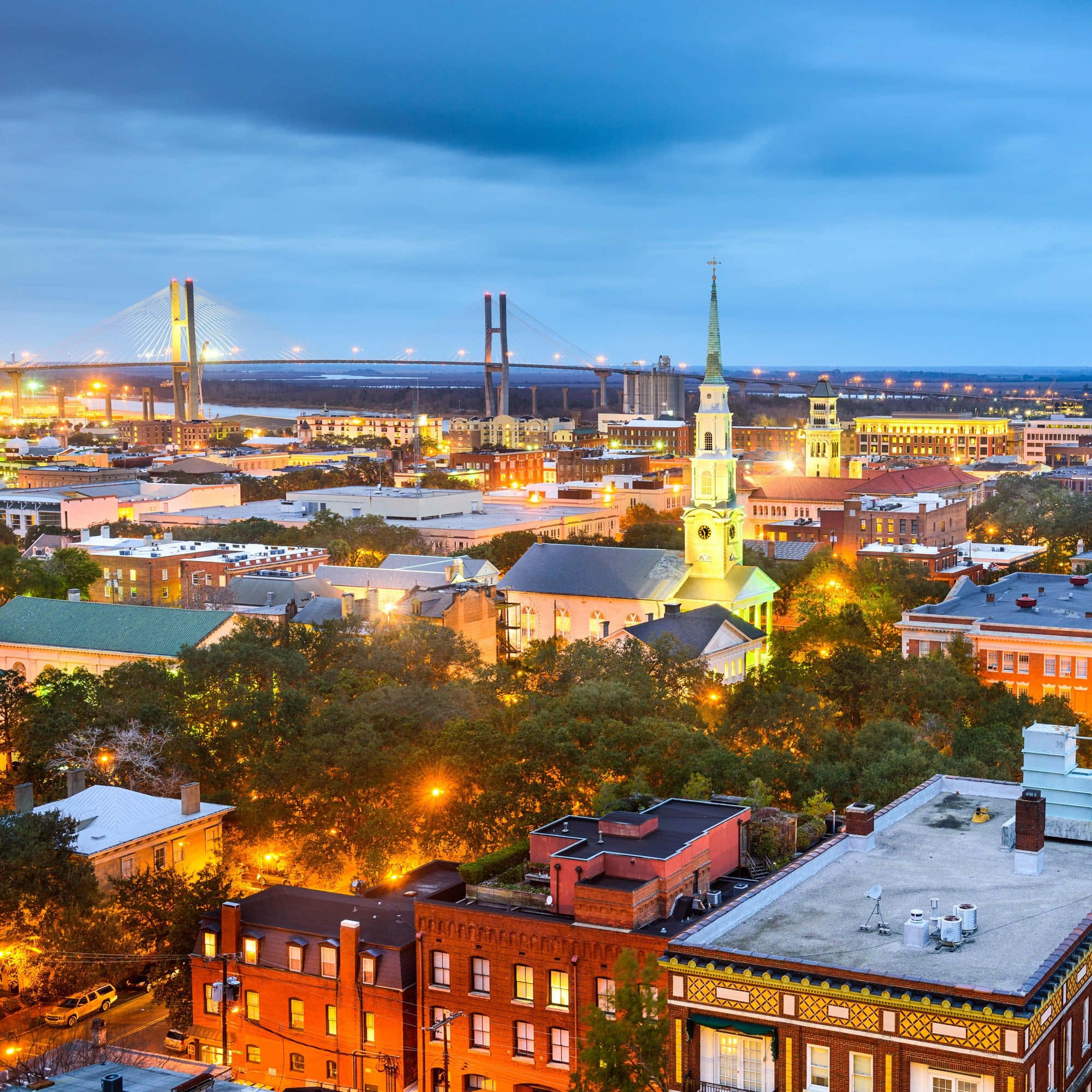 Savannah 桌布 壁紙 — 暮色城市景觀，建築物和樹木散發出溫暖的光芒，深藍色的天空下有一座遠方亮燈的橋樑。