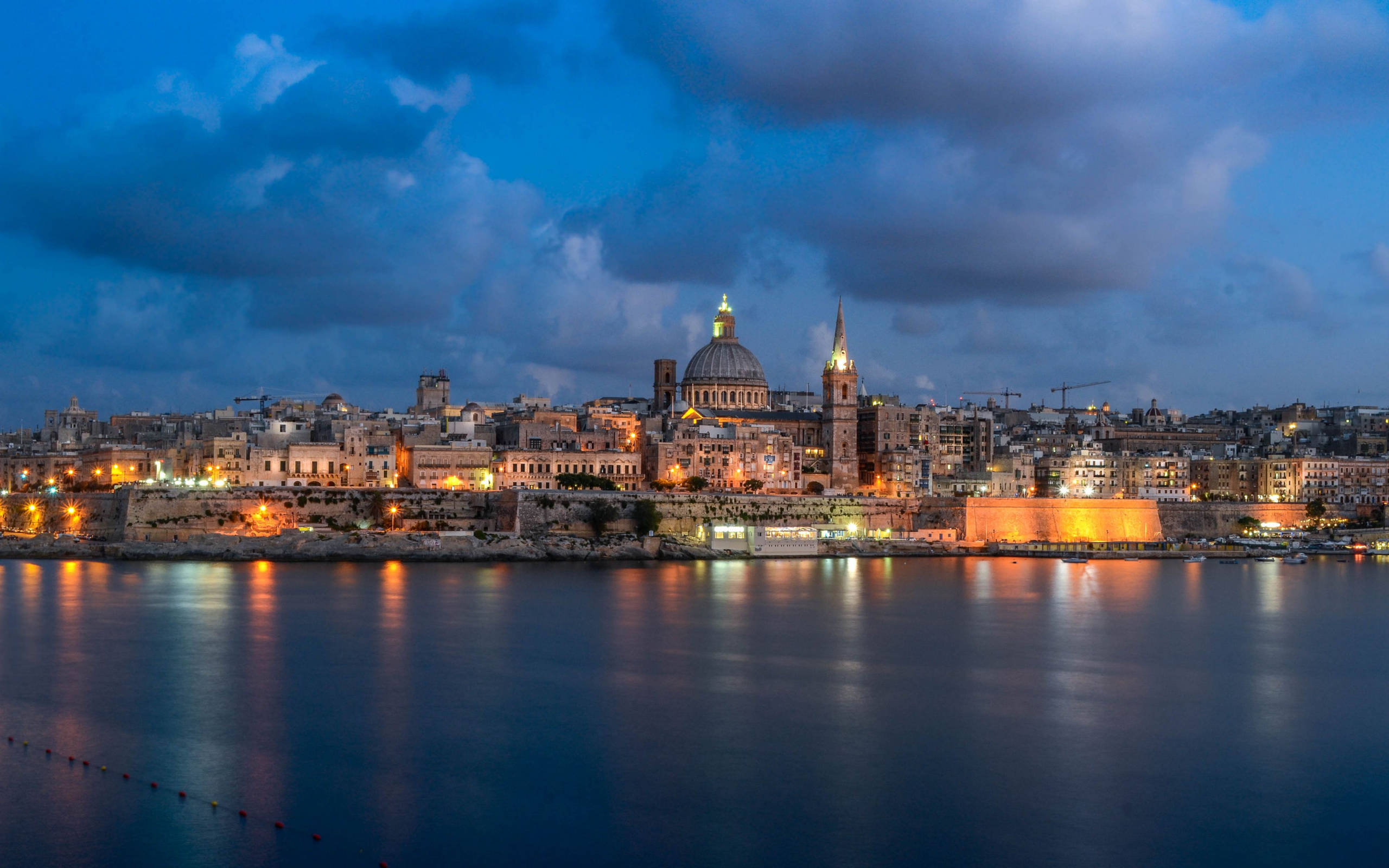 Malta 桌布 壁紙 — 暮色城市景觀，歷史悠久的圓頂建築和尖塔在憂鬱的藍天下溫暖地發光，倒映在平靜的水面上。