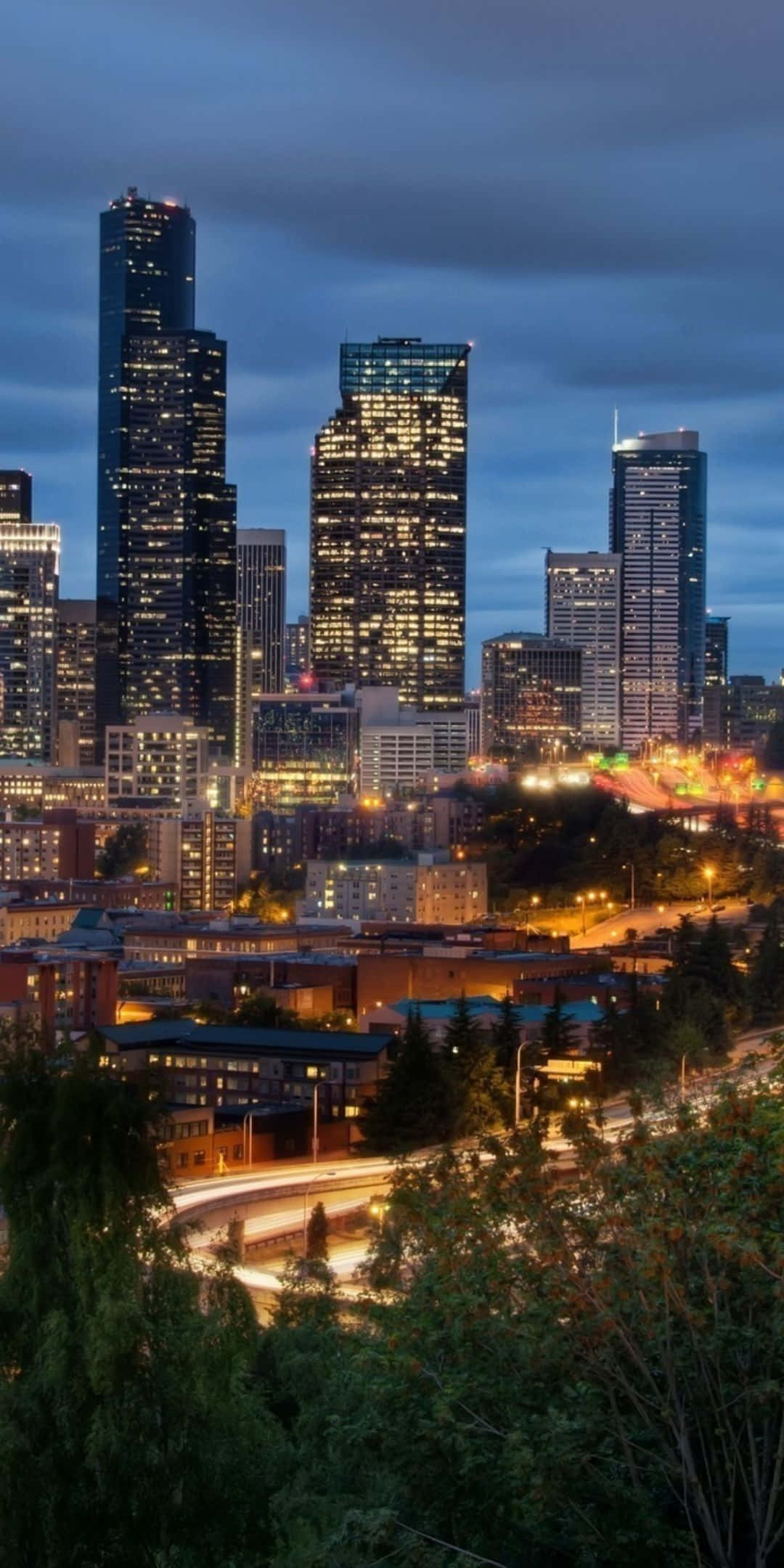 Pixel 3 Seattle 桌布 壁紙 — 暮色城市景觀，高聳的摩天大樓在昏暗的天空下閃耀，周圍環繞著茂密的綠樹。