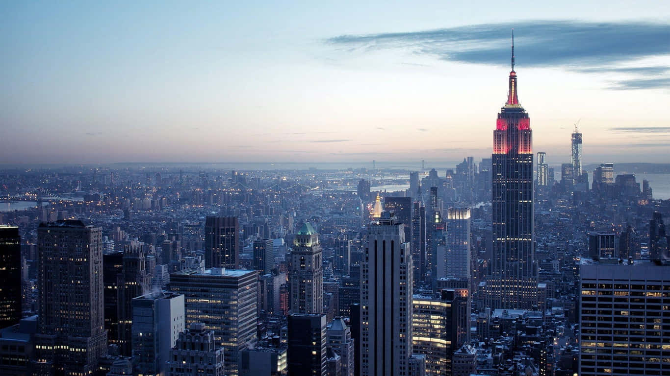 1366x768 Empire State Building 桌布 壁紙 — 暮色降臨紐約市，帝國大廈在柔和的藍天下閃耀著紅光。