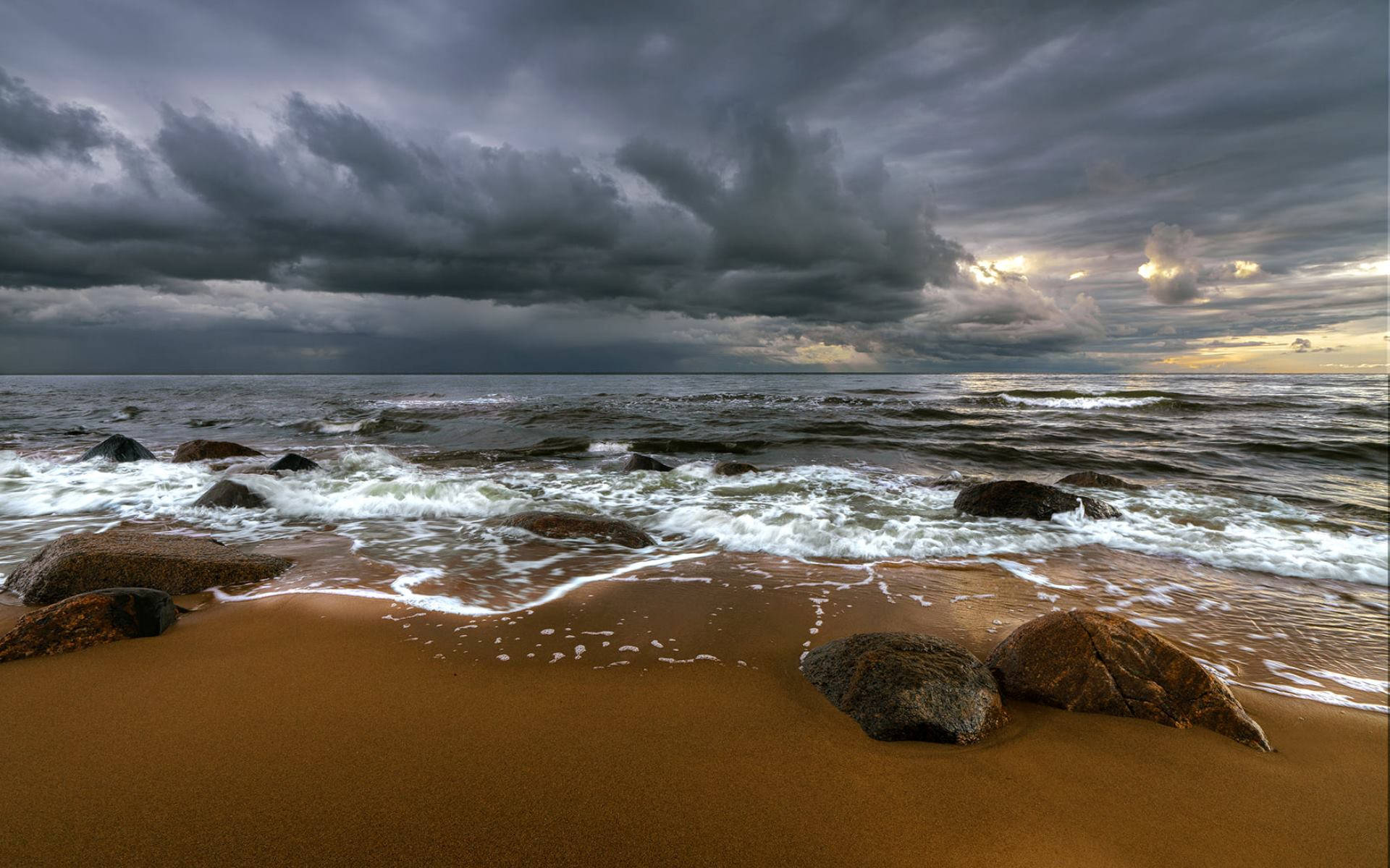 Latvia 桌布 壁紙 — 暴風雨的海浪拍打沙灘和暗黑岩石，天空烏雲密佈，帶有夕陽餘暉。