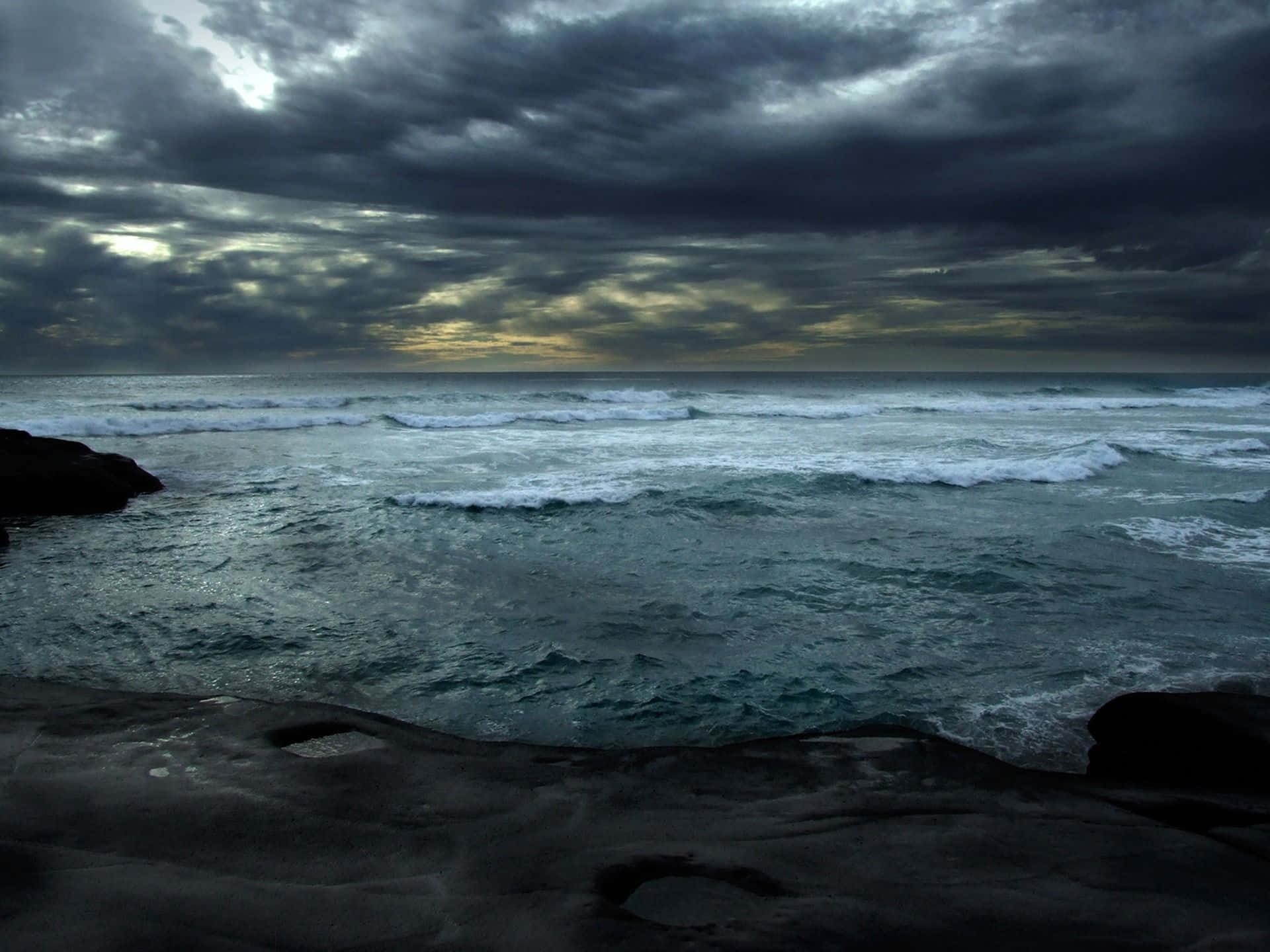 Dark Ocean 桌布 壁紙 — 暴風雨的海浪拍打著黑暗的岩石，在陰沉的暮色天空下，金色光芒隱約穿透雲層。