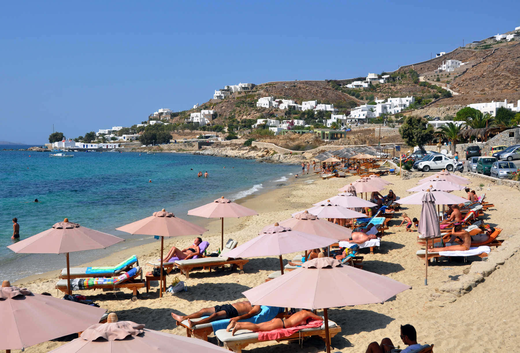 Greek Beach 桌布 壁紙 — 曬太陽的人們在沙灘上粉紅色遮陽傘下放鬆，沙灘旁有碧綠的海水和白色懸崖房屋，天空清澈湛藍。