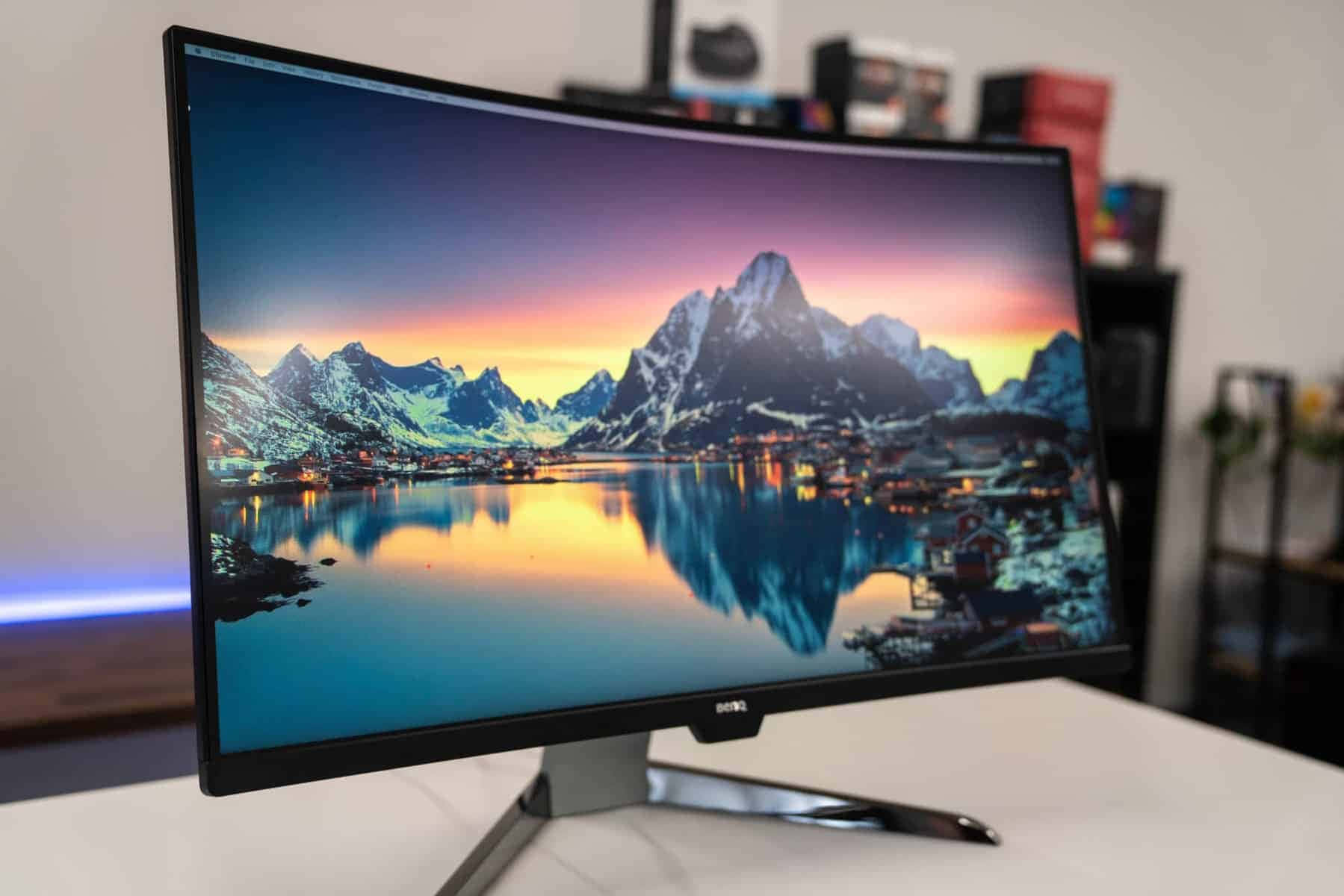 Gaming Monitors 桌布 壁紙 — 曲面顯示器展示了一個充滿活力的山湖日落，雪覆蓋的山峰和發光的村莊燈光倒映在平靜的水面上。