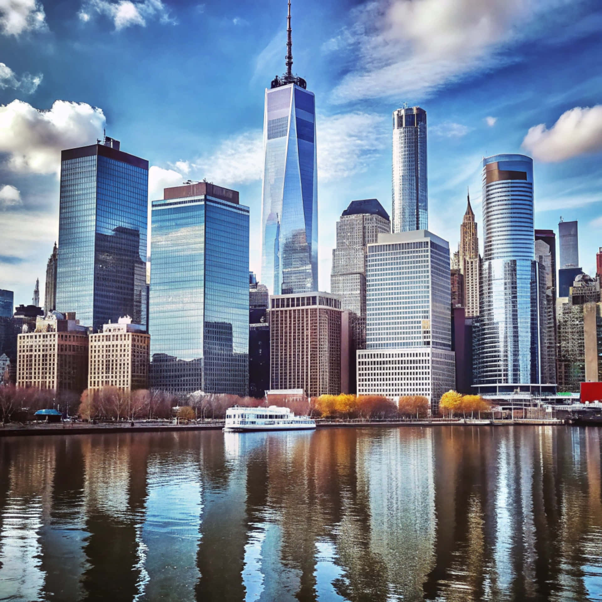 New York Skyline 壁紙 — 曼哈頓下城高聳的玻璃摩天大樓倒映在水中，一艘白色小船在秋樹旁滑過，天空湛藍。