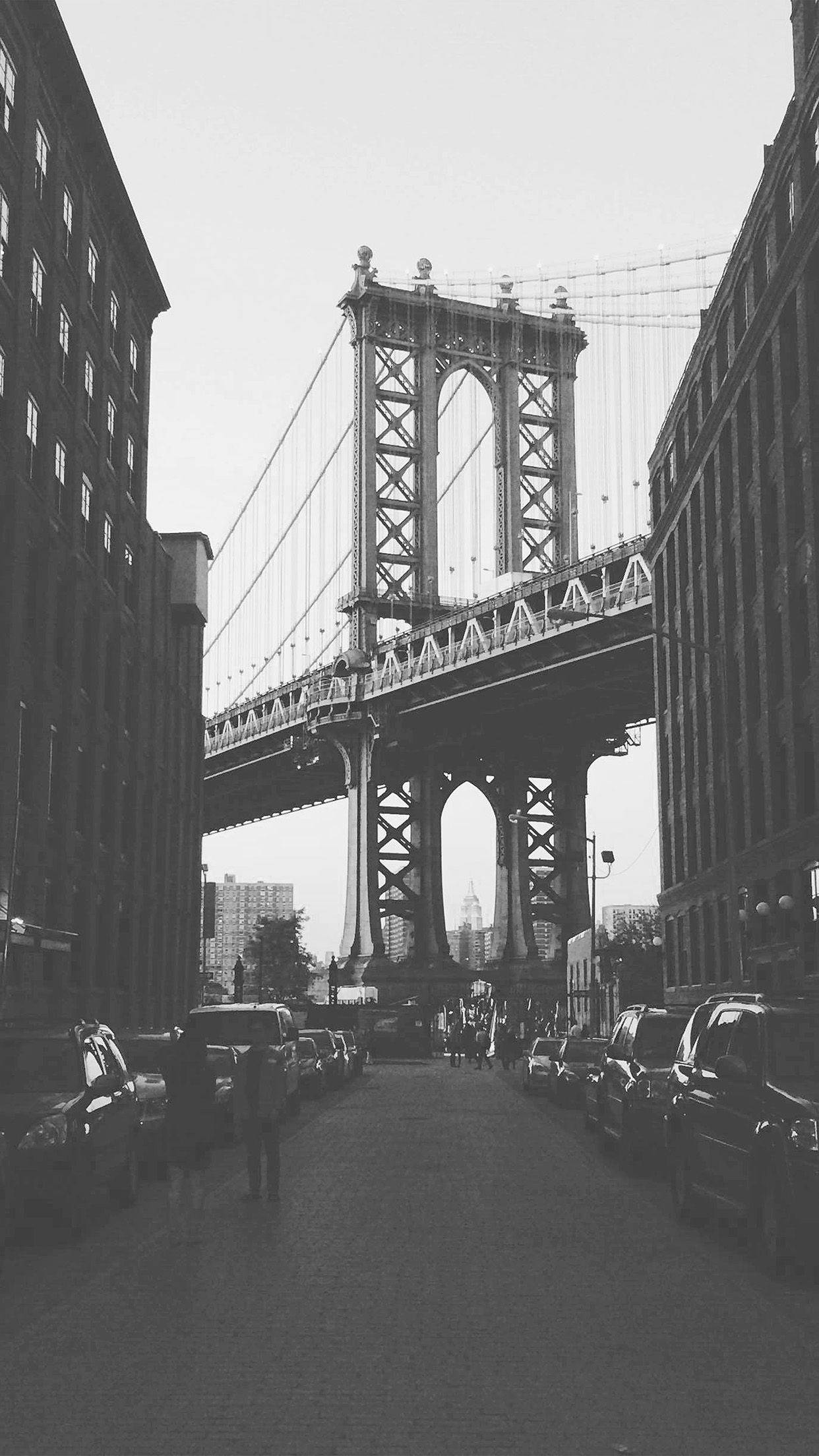 Black And White iPhone 桌布 壁紙 — 曼哈頓大橋的黑白景觀，聳立在一條狹窄的城市街道上，兩旁停滿汽車和高樓大廈。