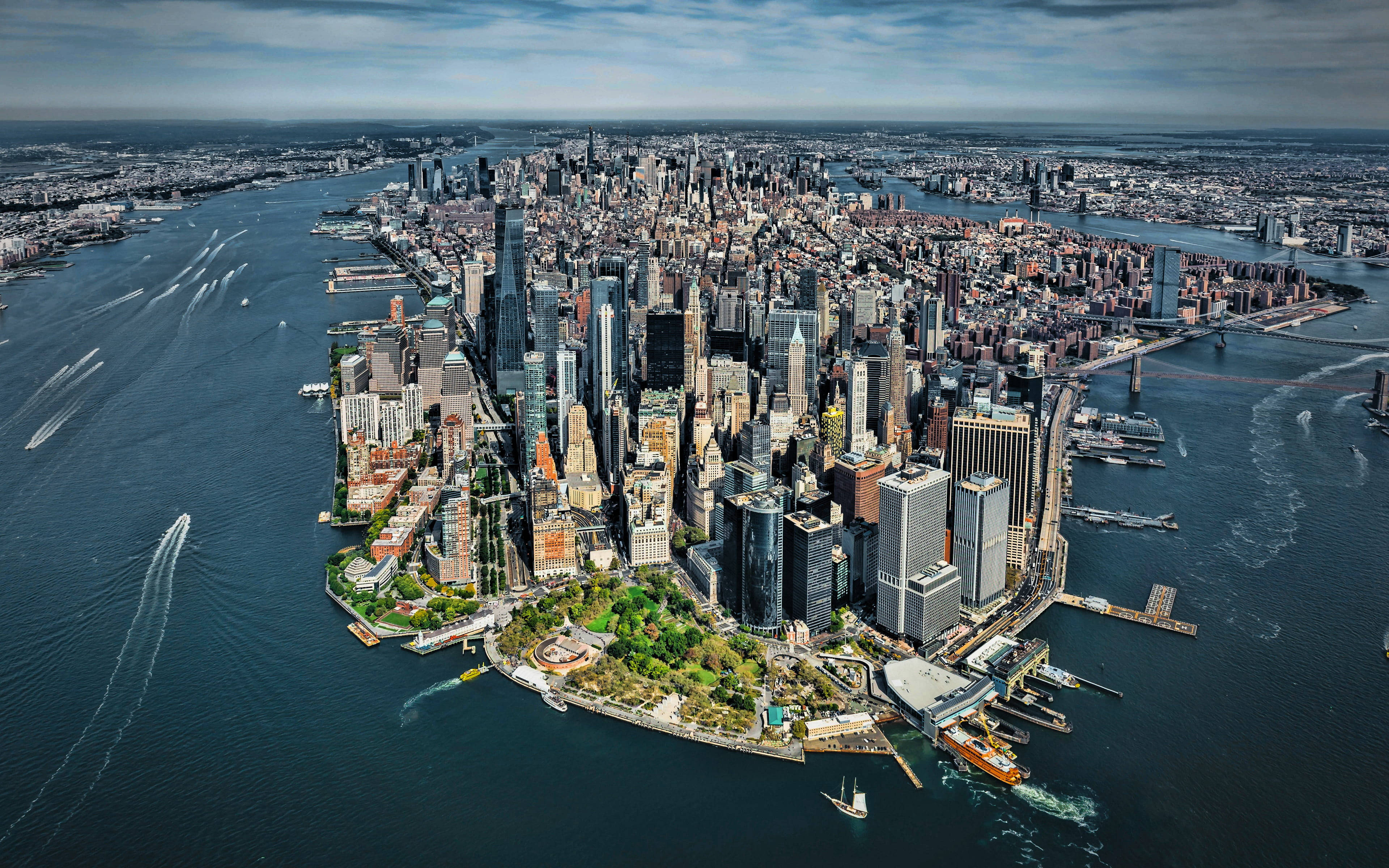 New York 桌布 壁紙 — 曼哈頓密集摩天大樓和河畔碼頭的鳥瞰圖，天空多雲，船隻留下白色尾跡。