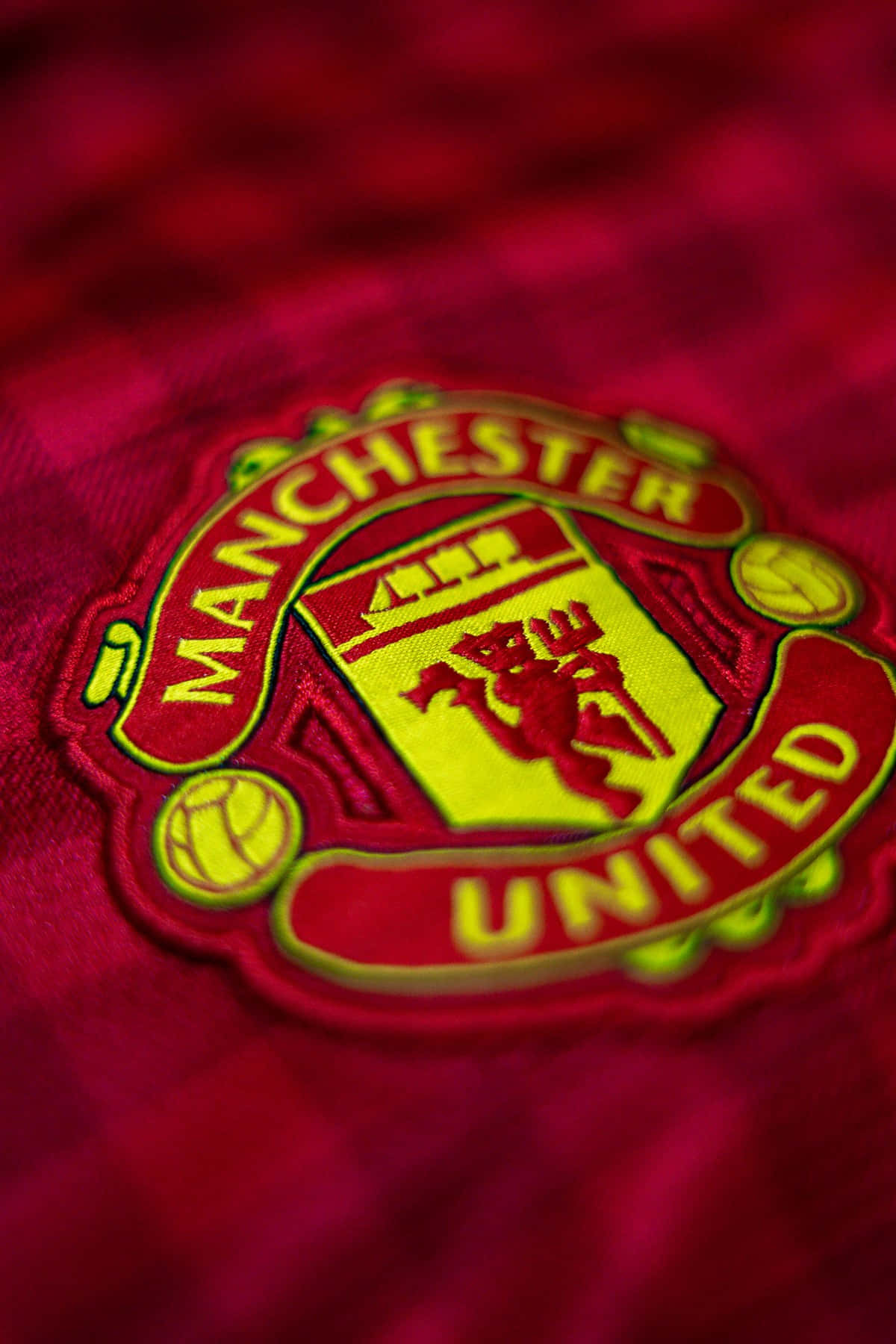 Manchester United iPhone 桌布 壁紙 — 曼聯隊徽近距離特寫，以紅色和金色刺繡在帶有紋理的紅色布料背景上。