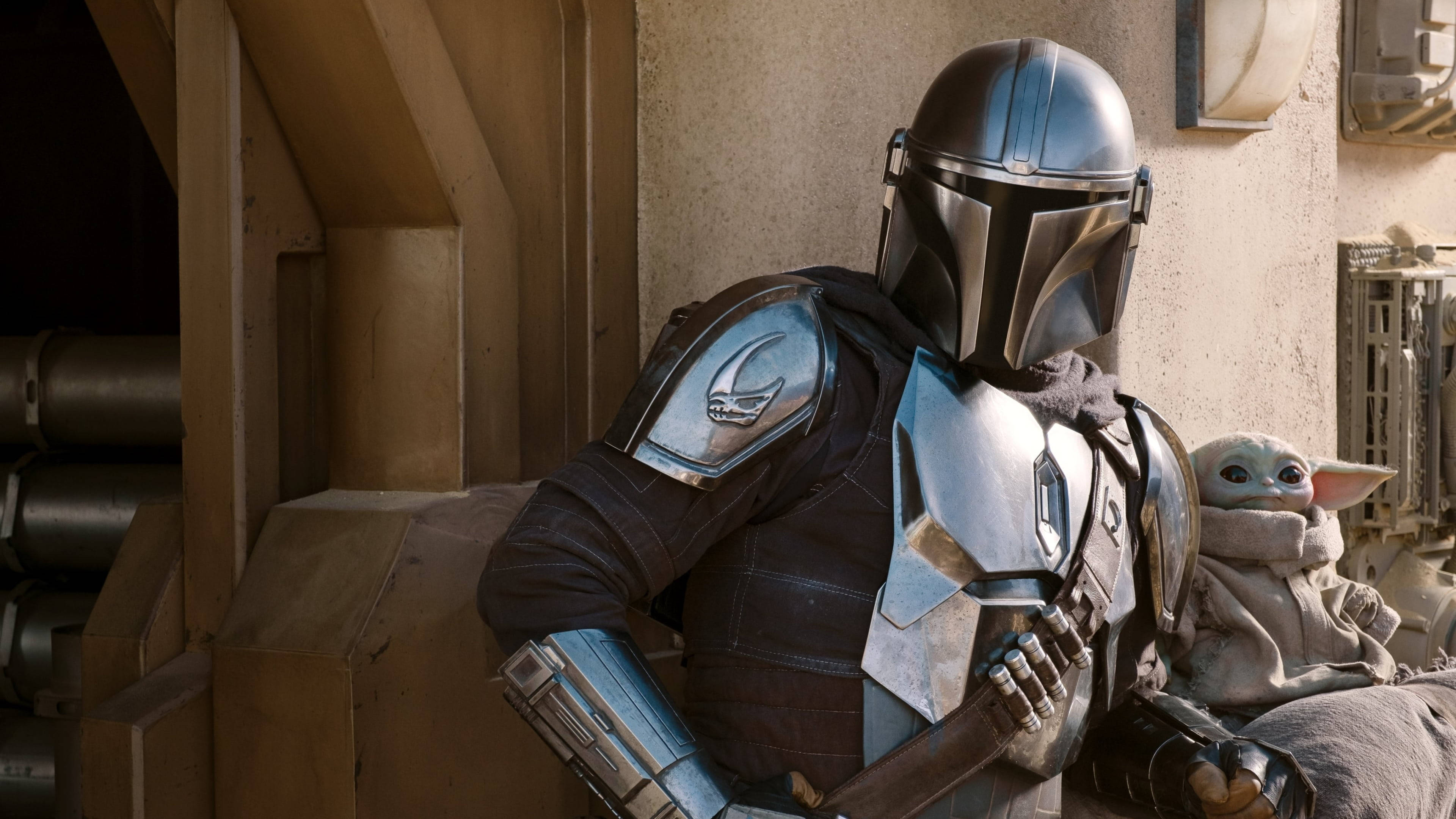 Mandalorian And Baby Yoda 桌布 壁紙 — 曼達洛人穿著他標誌性的銀色盔甲，在一個粗獷、溫暖照明的太空船內部抱著孩子古古。