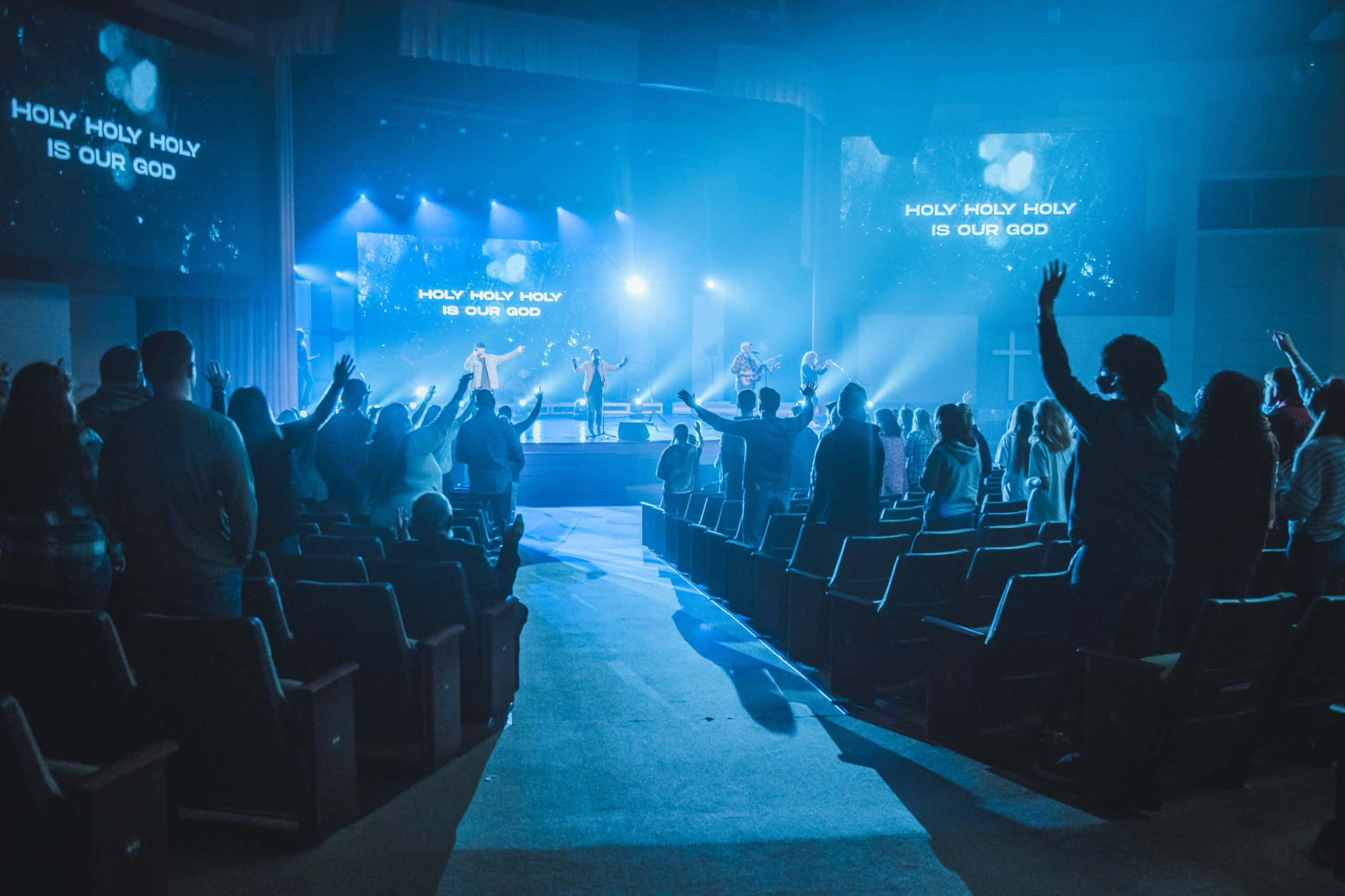 Church Stage 桌布 壁紙 — 會眾在燈光昏暗的教堂中敬拜，藍色舞台燈光下，音樂家在升高平台上表演，會眾舉手敬拜。