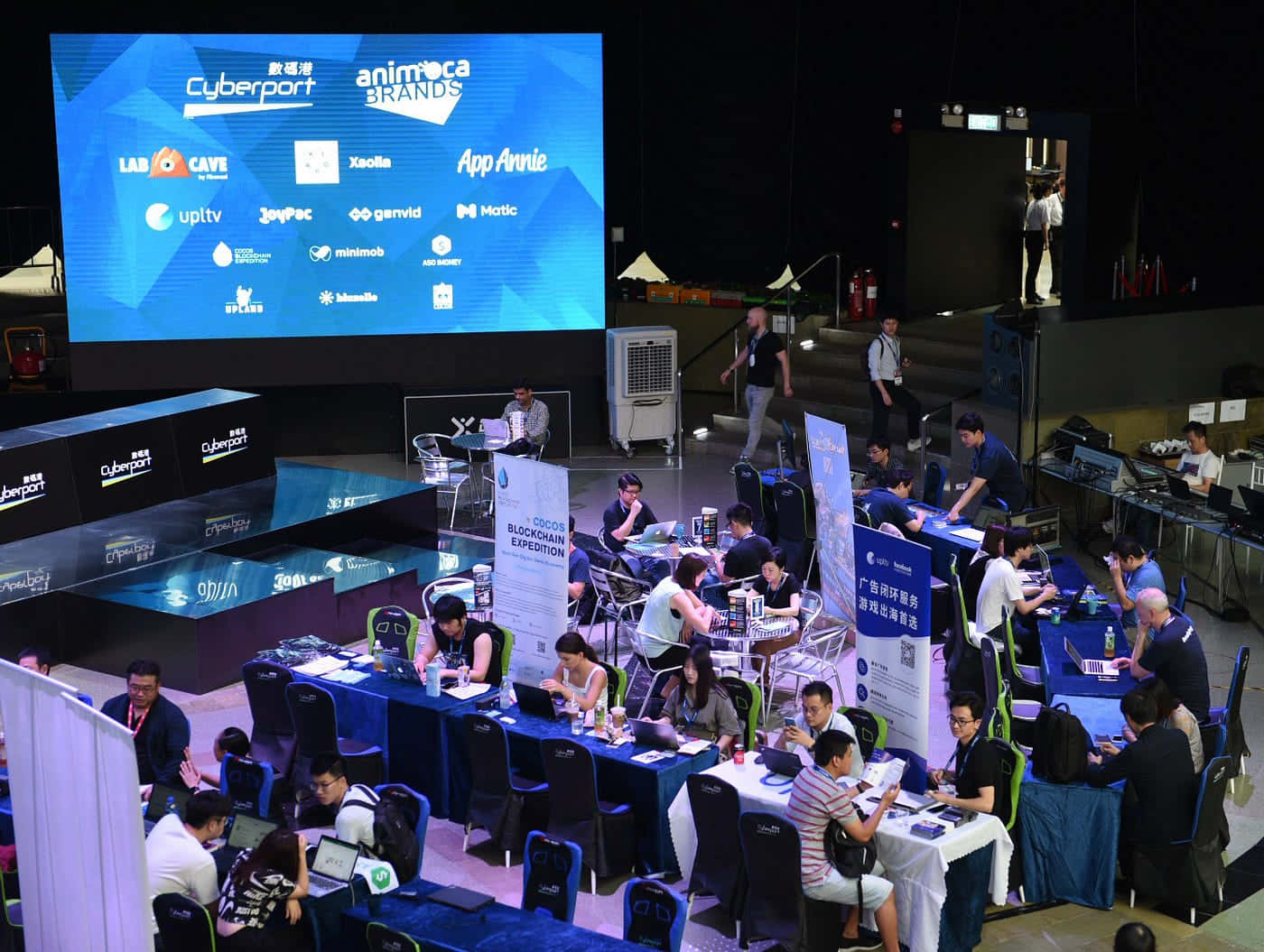 Gaming Conventions 桌布 壁紙 — 會議廳內，一大群與會者在展示科技標誌的大型螢幕下的攤位使用筆記型電腦工作。