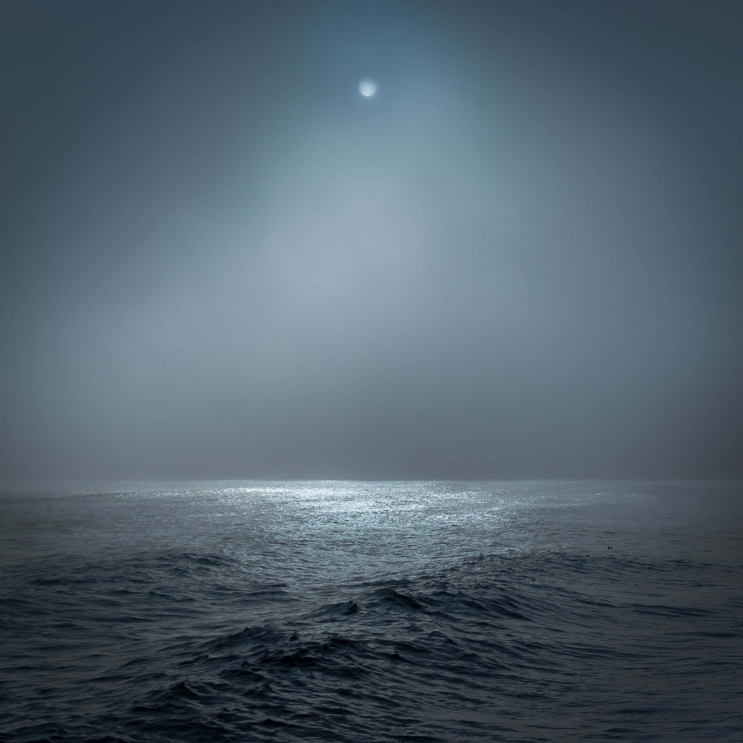 Dark Ocean 桌布 壁紙 — 月光在朦朧的夜空中閃爍，映照在霧氣瀰漫的海洋上，帶有柔和的藍色和灰色調。