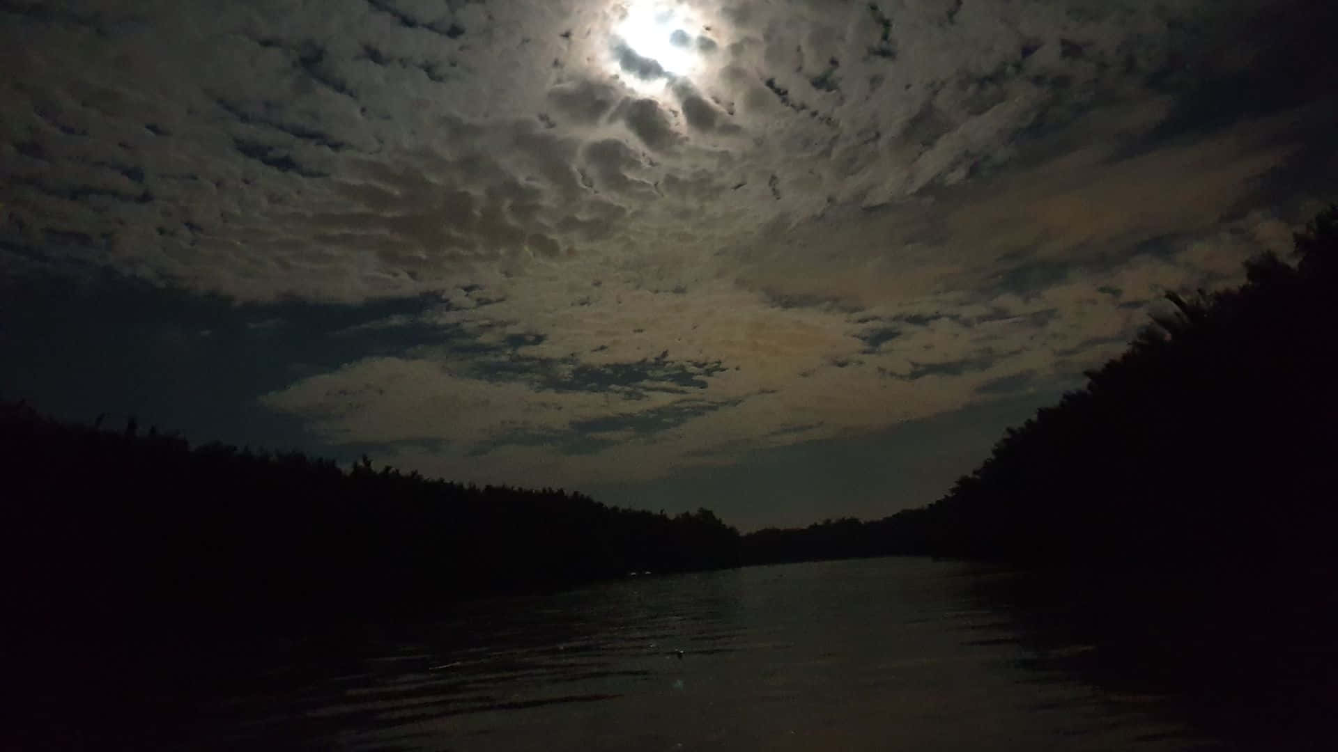 黑夜 桌布 壁紙 — 月光穿透有紋理的雲層，照耀在兩旁被茂密森林剪影環繞的黑暗河流上。