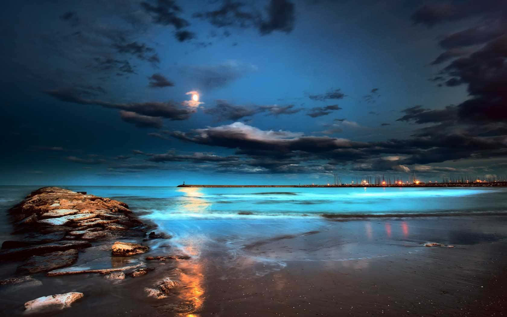 海灘 桌布 壁紙 — 月光穿過雲層，照耀在寧靜的海灘上，閃爍著碧綠色的波浪和地平線上遙遠的城市燈光。