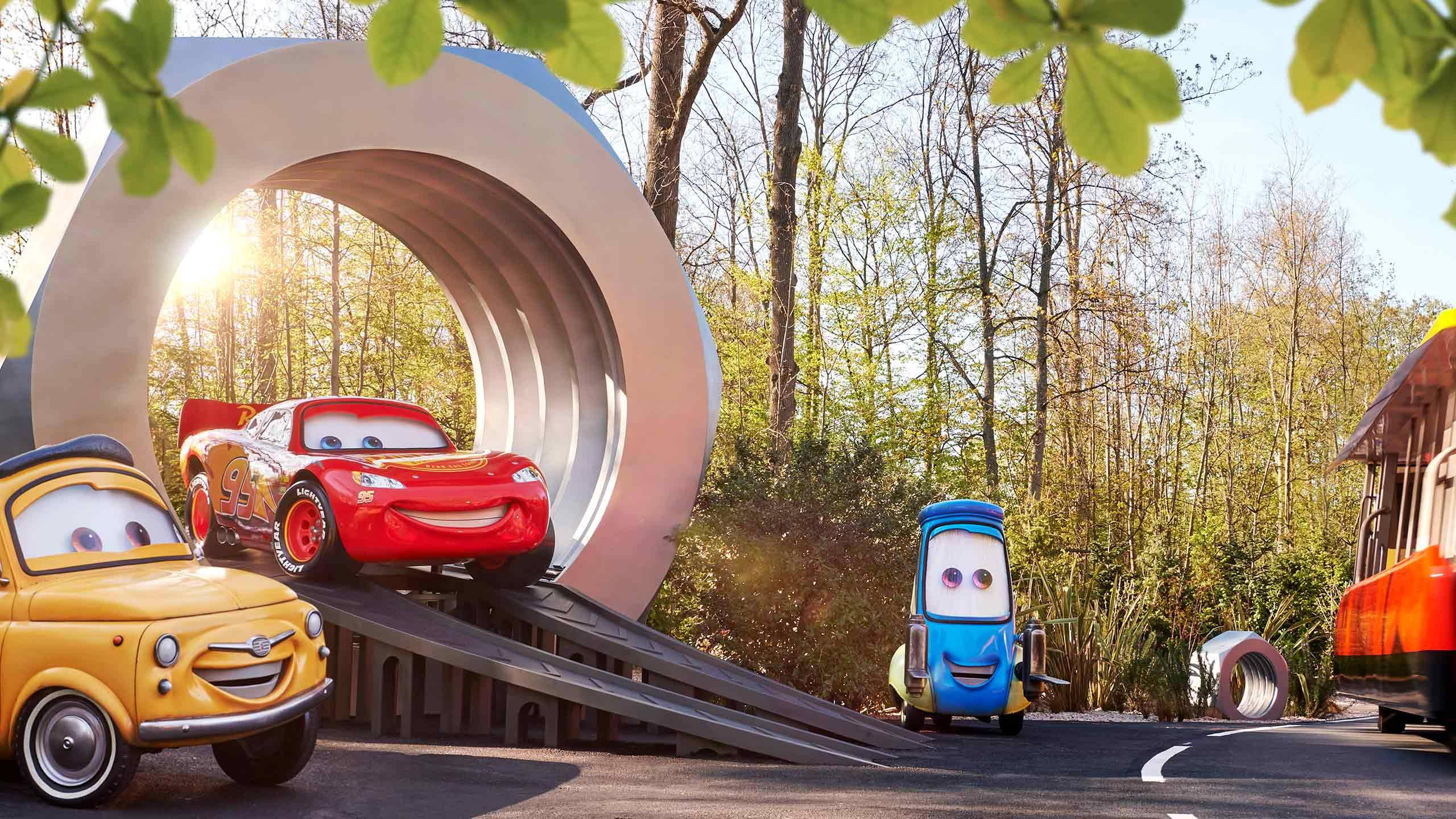 迪士尼汽車總動員 桌布 壁紙 — 有面孔的動畫汽車在陽光明媚的森林中歡快地聚集在一個巨大的金屬輪胎坡道周圍。
