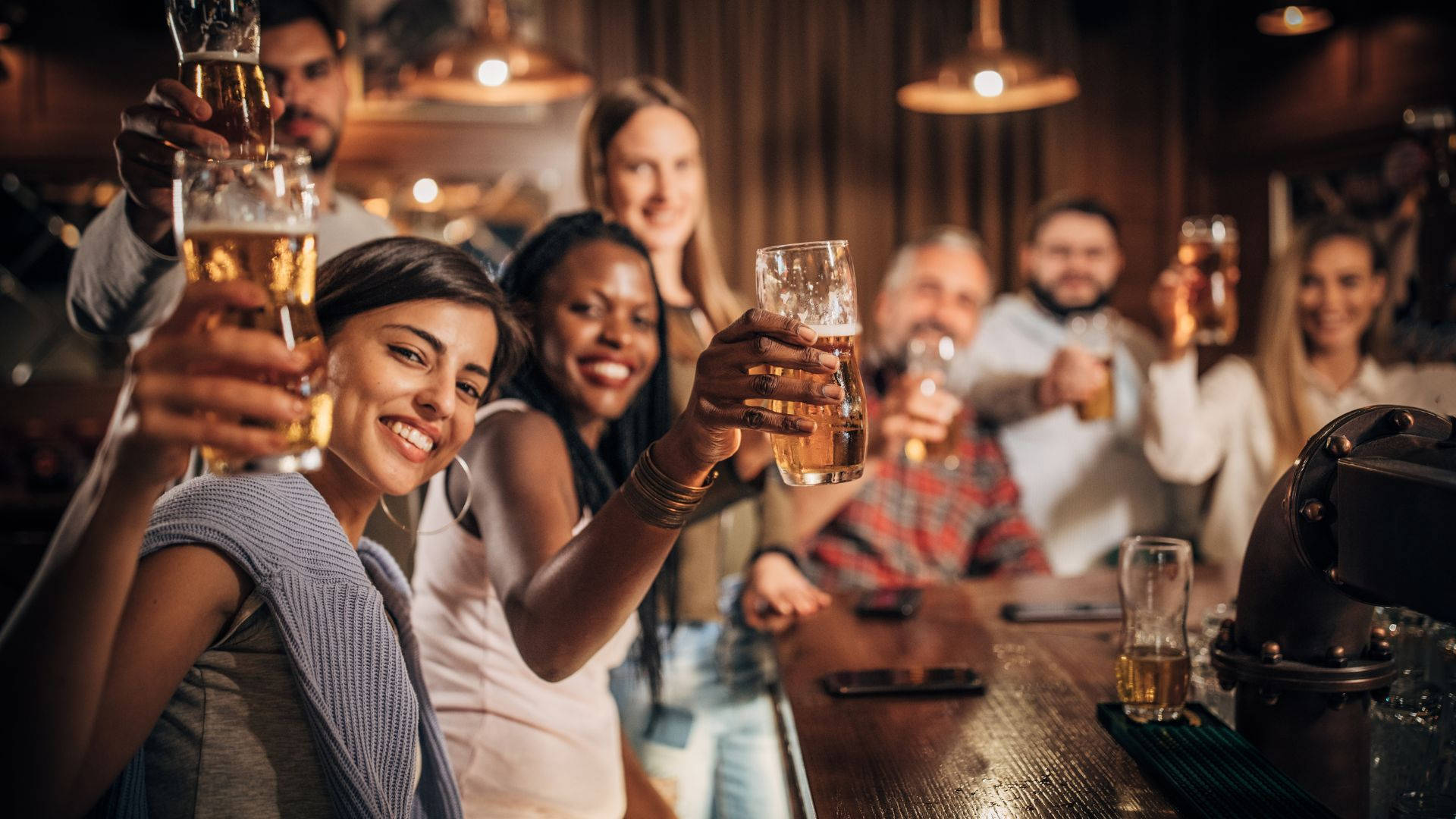drinks 桌布 壁紙 — 朋友們在吊燈下溫暖照亮的酒吧櫃檯前，舉著啤酒杯歡呼。
