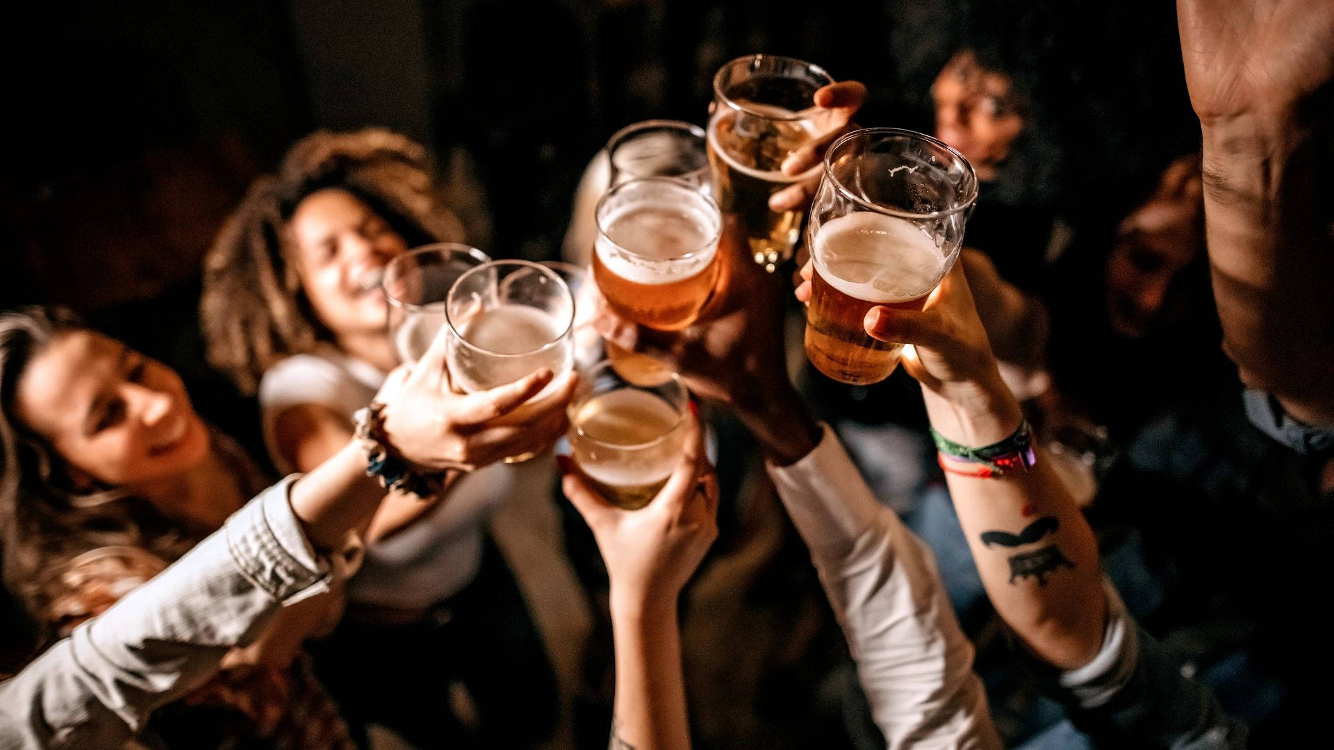 drinks 桌布 壁紙 — 朋友們在溫暖昏暗的燈光下舉起琥珀色啤酒杯，在熱鬧的派對上乾杯。
