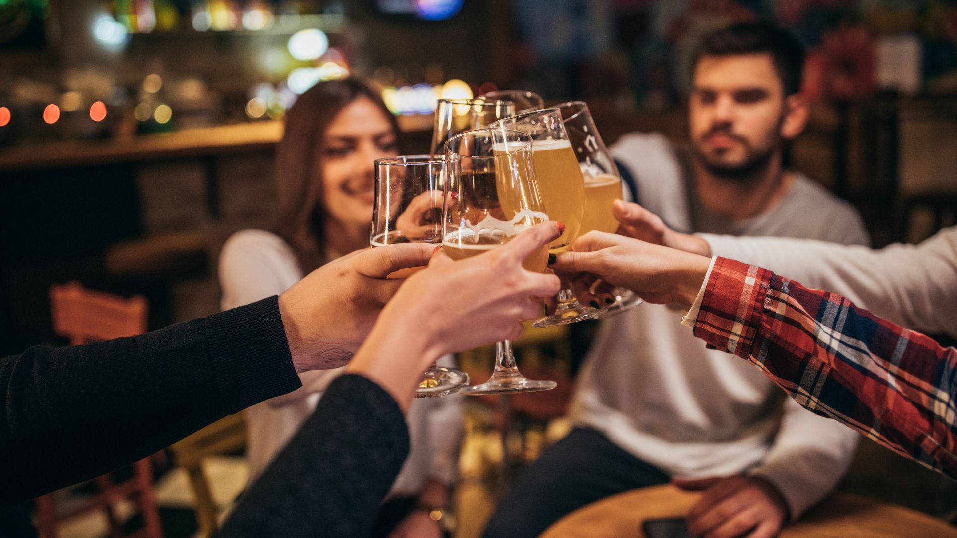 drinks 桌布 壁紙 — 朋友們在溫暖燈光的酒吧中碰啤酒杯，背景燈光模糊，臉帶微笑。