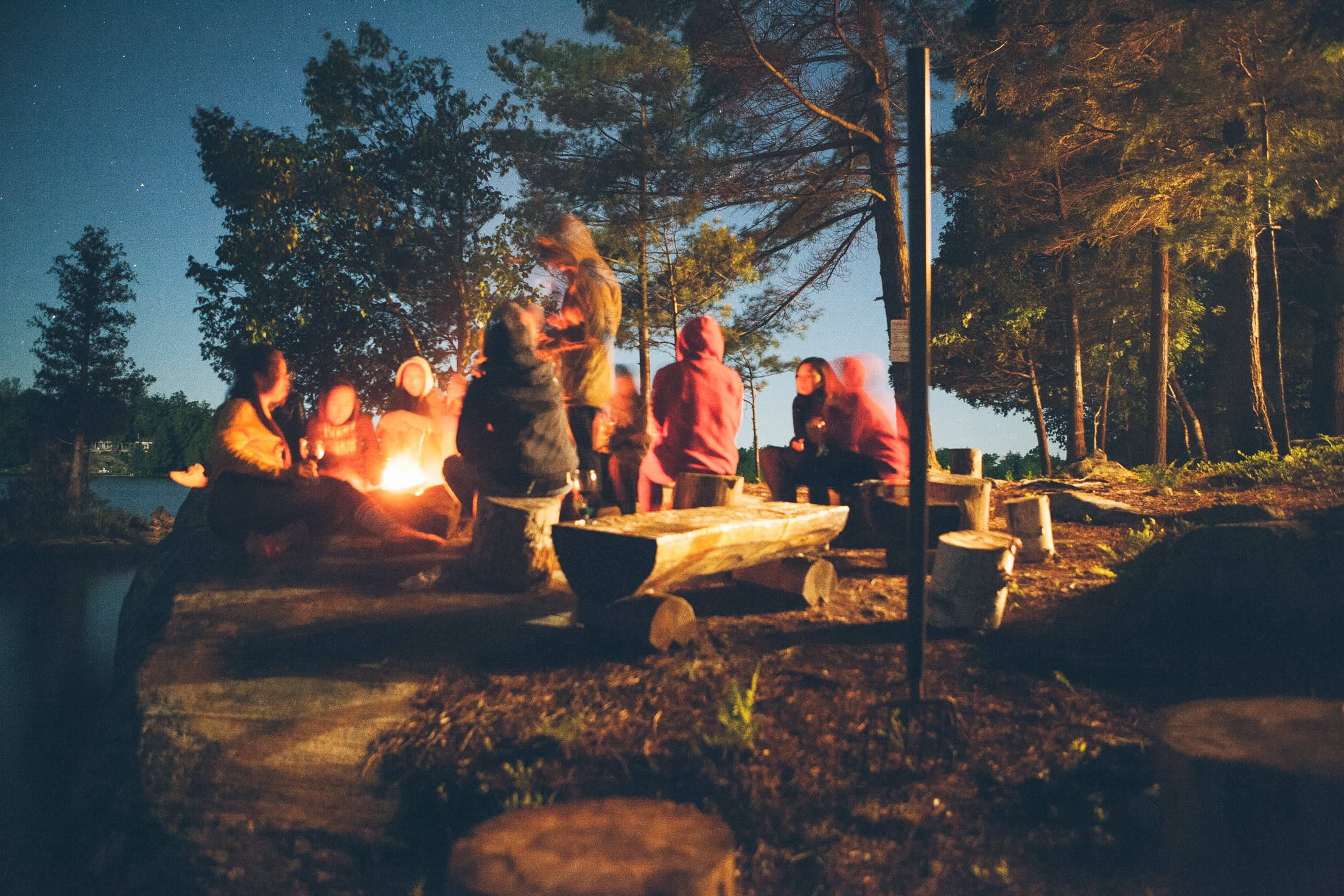 Camping 桌布 壁紙 — 朋友們在黃昏時分聚集在森林中一個寧靜的湖邊，在星空下圍著發光的營火。