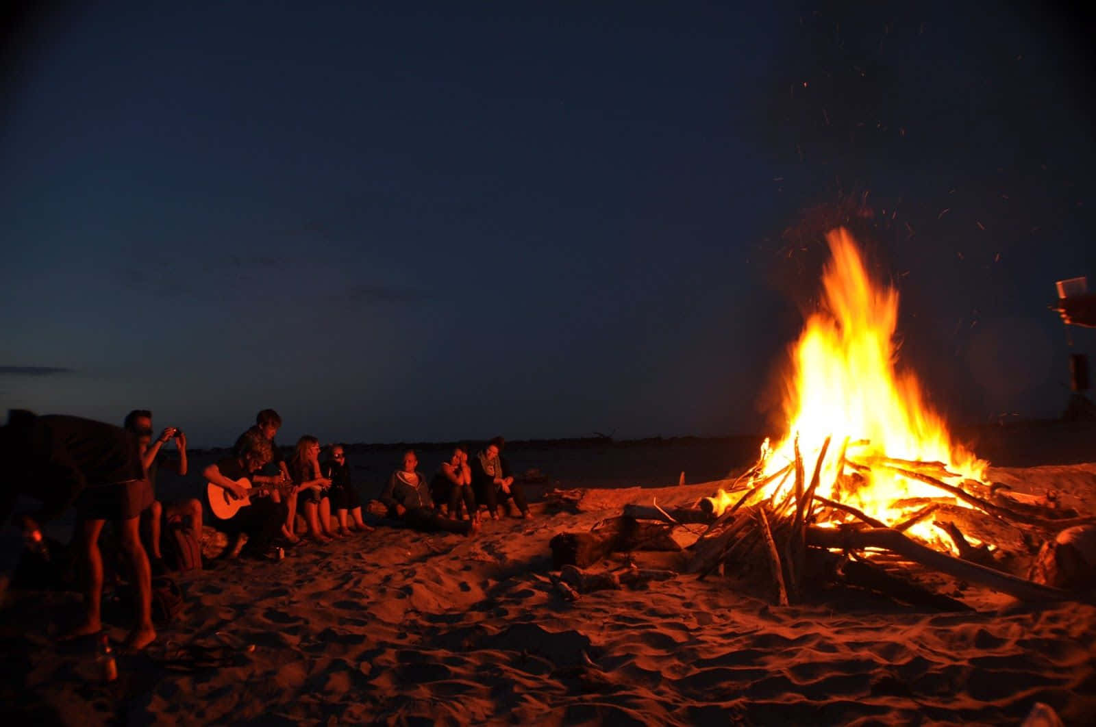 Beach Camping 桌布 壁紙 — 朋友們在黃昏時分聚集在熊熊燃燒的海灘營火旁，溫暖的橙色光芒灑在沙灘和剪影上。