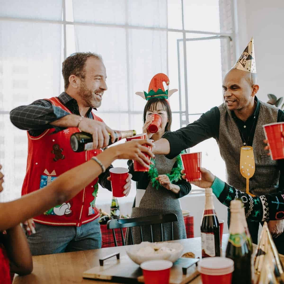 聖誕節 桌布 壁紙 — 朋友們穿著節日毛衣和派對帽，圍著擺滿飲料和小吃的桌子，高舉紅色杯子歡呼。