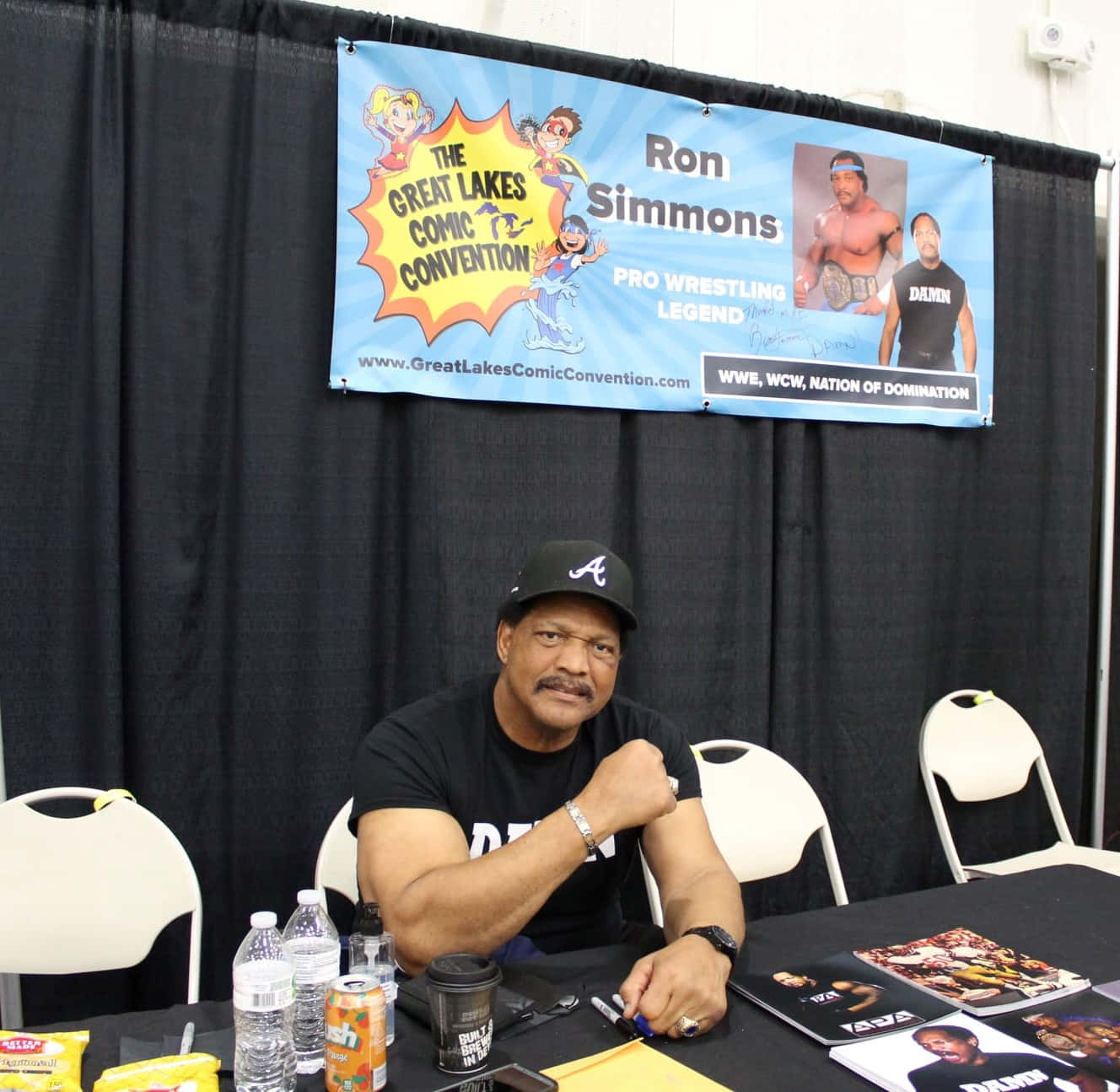 WWE 桌布 壁紙 — 朗·西蒙斯坐在簽名桌後，上方掛著Great Lakes Comic Convention的橫幅，宣傳他的職業摔角傳承。