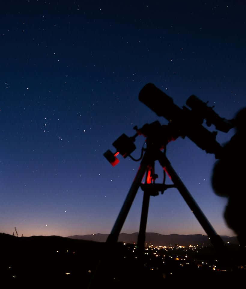 astronomy 桌布 壁紙 — 望遠鏡安裝在三腳架上，指向遠方城市燈光與山丘之上的星空剪影。