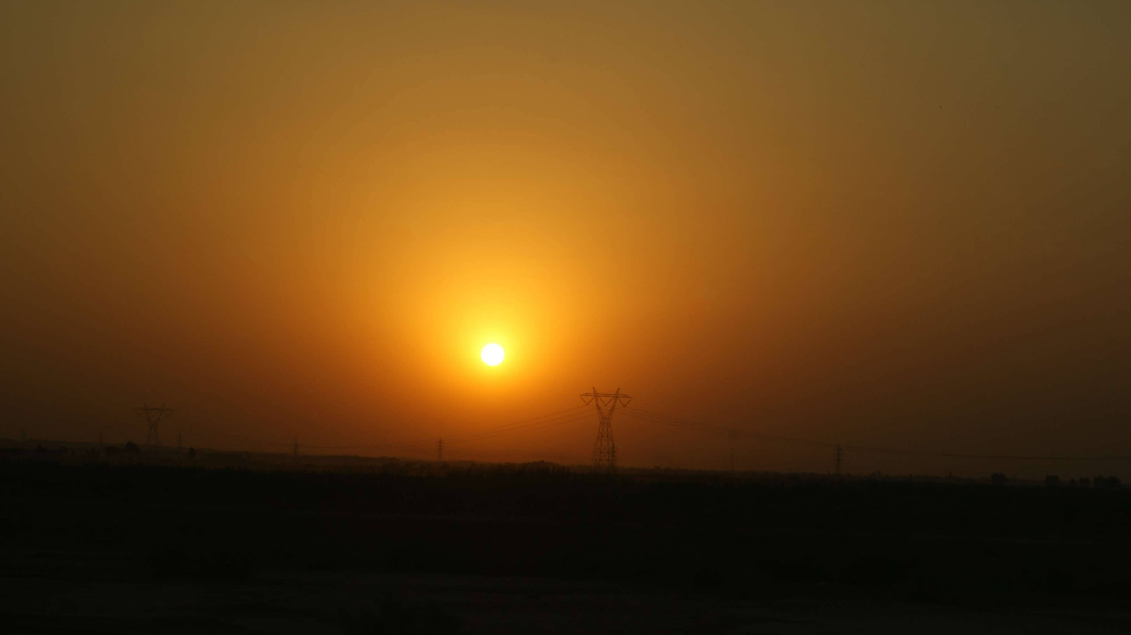 Iraq 桌布 壁紙 — 朦朧的日落將溫暖的橙色光芒投射在平坦的景觀上，襯托出電線和電塔的剪影。