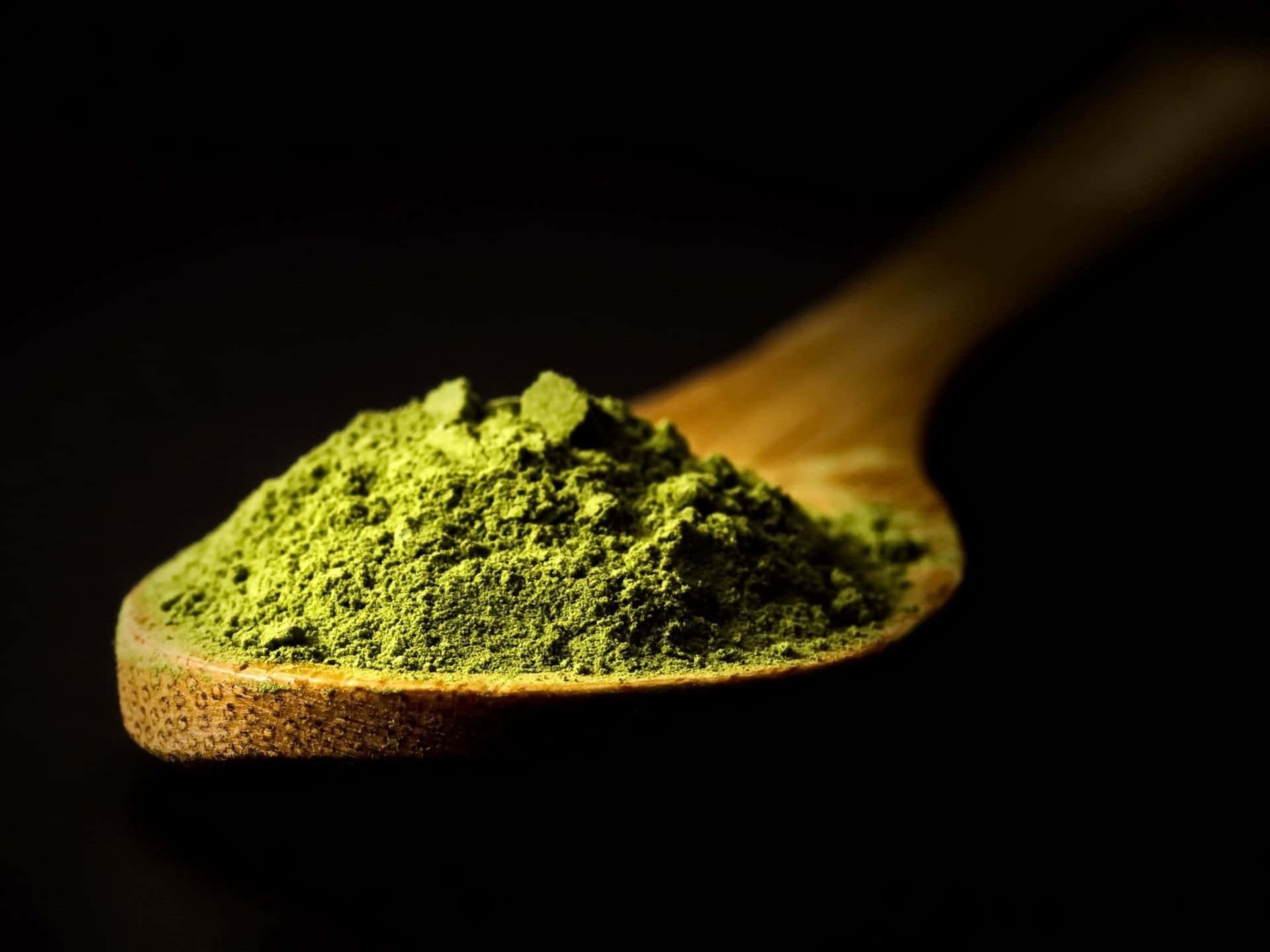 Matcha 桌布 壁紙 — 木勺盛著鮮綠色的抹茶粉，在黑色背景上呈現戲劇性的燈光效果。