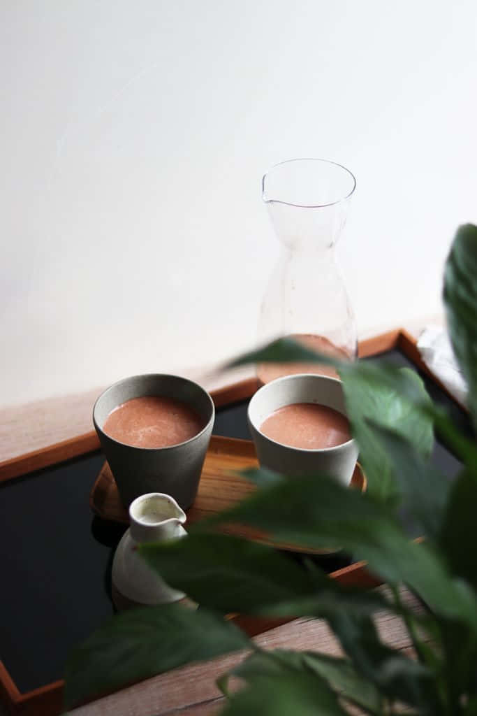 Minimalist Food 桌布 壁紙 — 木托盤上兩個陶瓷杯裝有粉紅色飲料，前景有玻璃水瓶和綠色植物葉子。