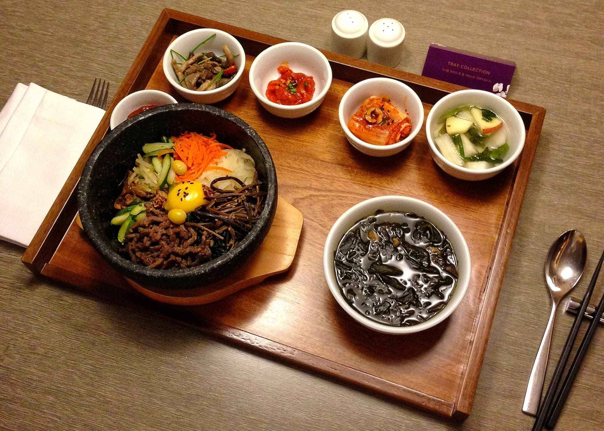 烹飪 桌布 壁紙 — 木托盤上放著一個滋滋作響的石鍋拌飯，配有色彩繽紛的蔬菜和牛肉，周圍有配菜和湯。
