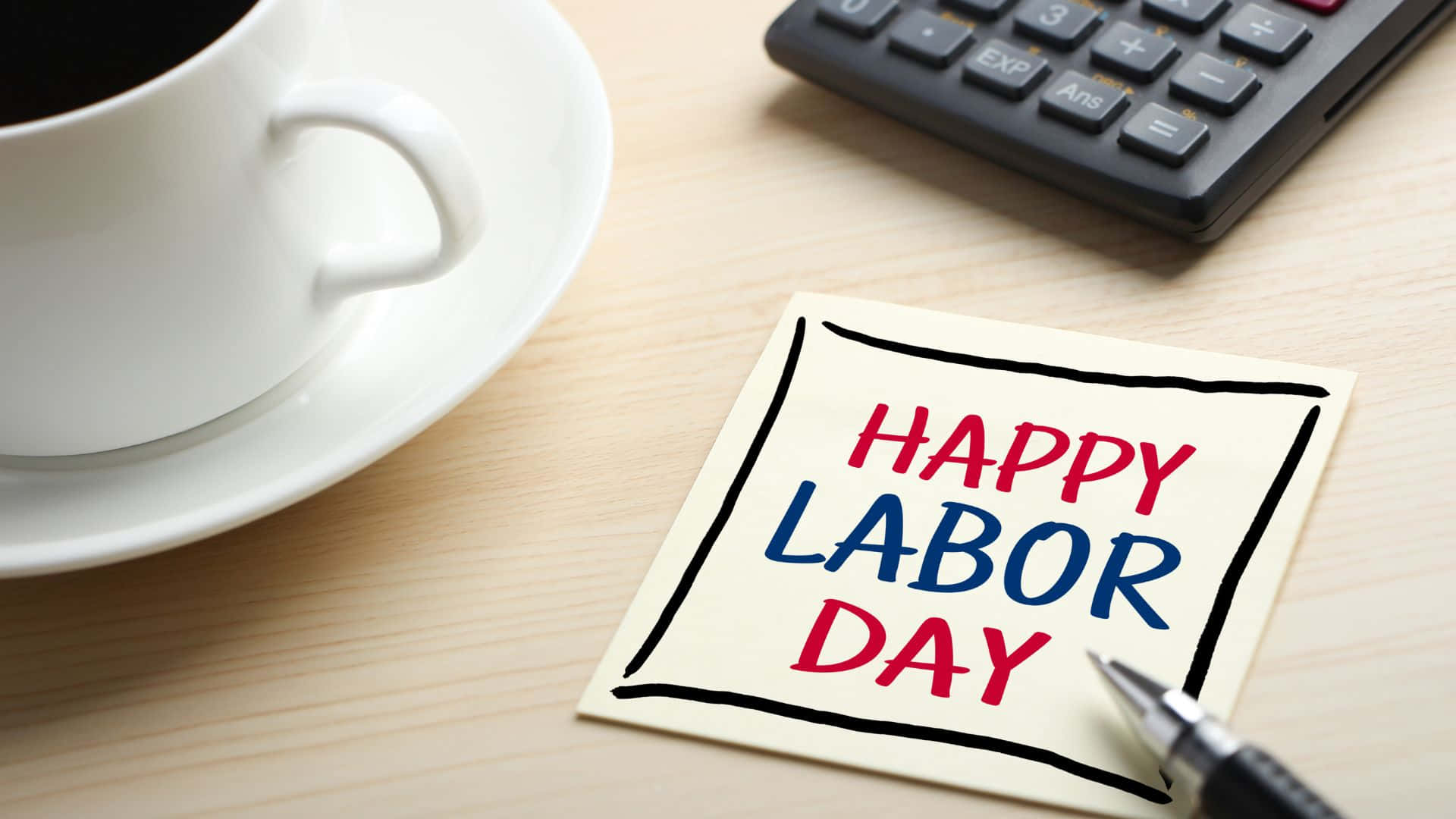 勞動節 桌布 壁紙 — 木桌上，咖啡杯、計算機和筆旁邊有一張手寫的「Happy Labor Day」便條。