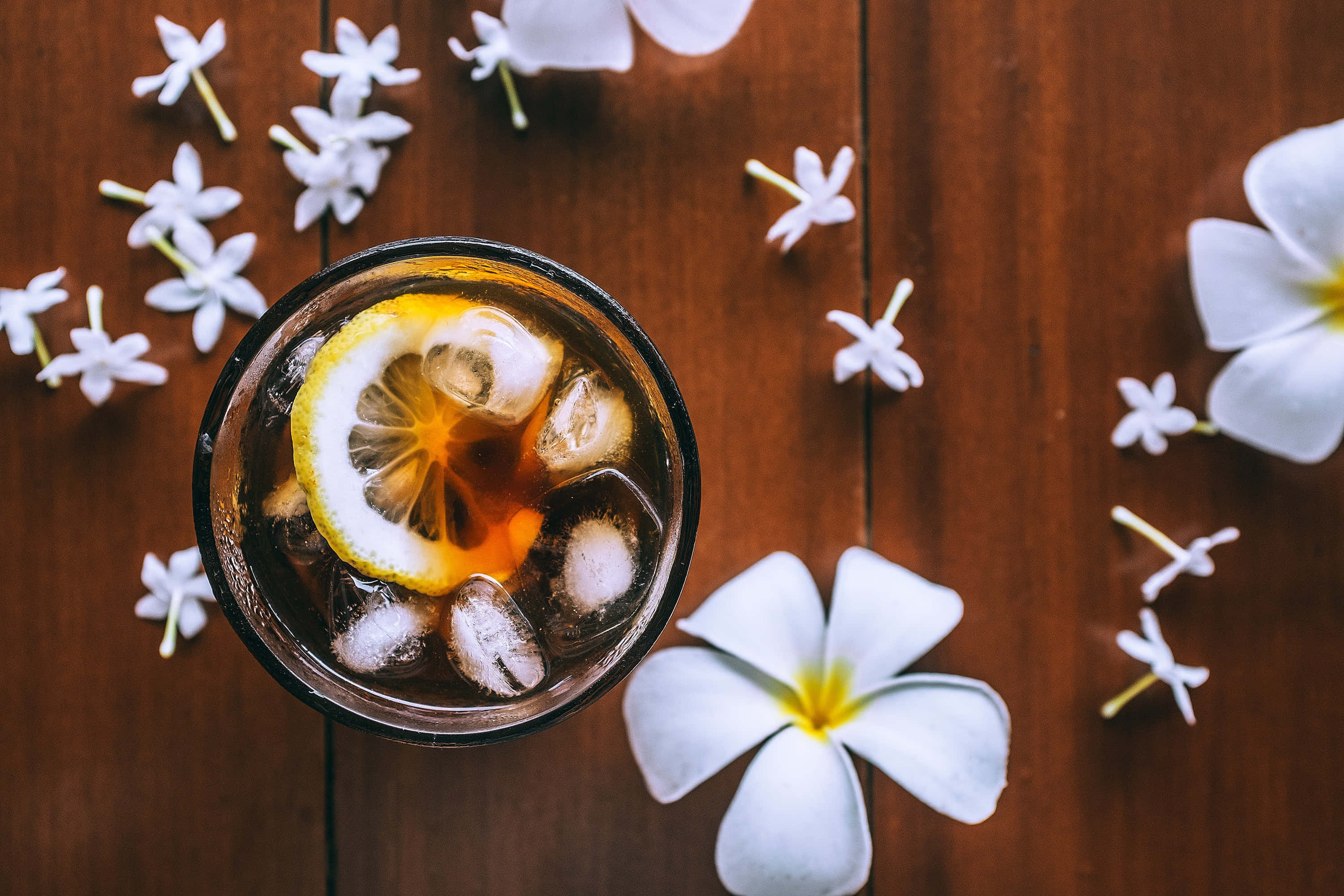 Summer Drinks 桌布 壁紙 — 木桌上深琥珀色雞尾酒配冰塊和檸檬片，周圍環繞白色雞蛋花，俯視角度。