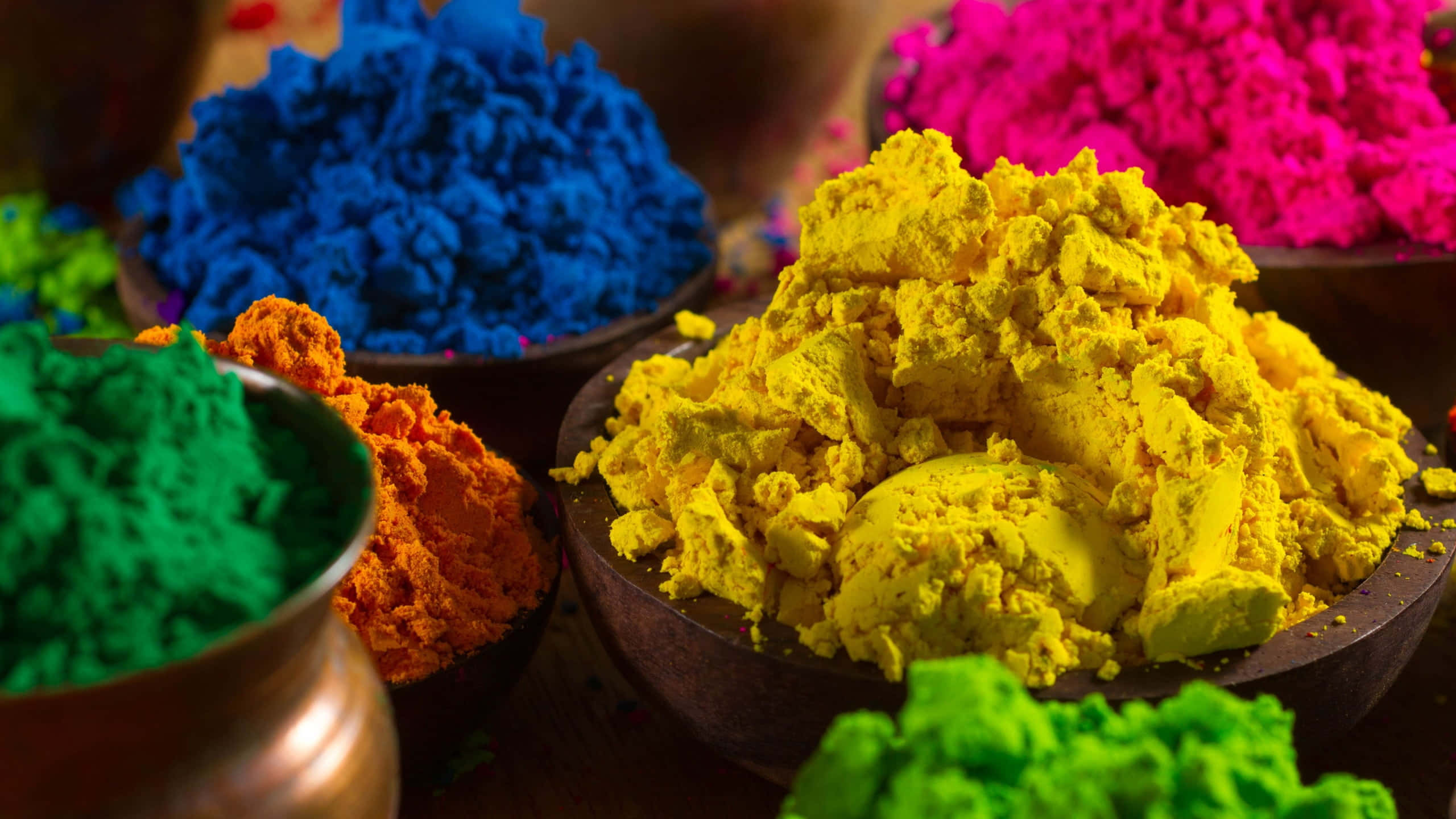 Holi 桌布 壁紙 — 木碗中色彩繽紛的粉末堆在溫暖燈光下閃耀，捕捉了侯麗節的節日氣氛。