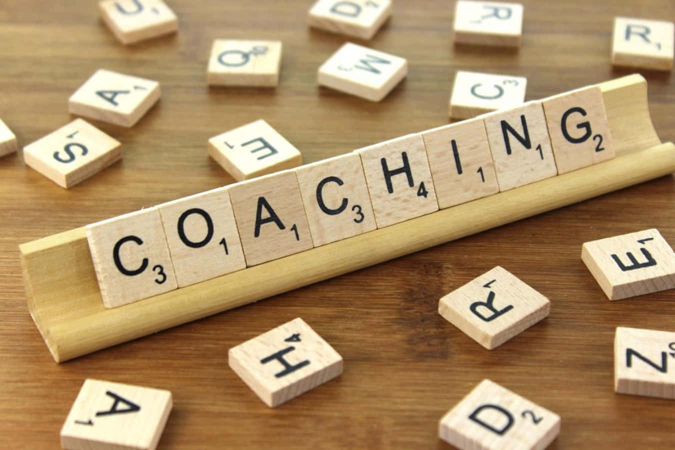Coach 桌布 壁紙 — 木製 Scrabble 字母牌在木架上拼出 COACHING，置於溫暖的木桌上，周圍散落著字母牌。