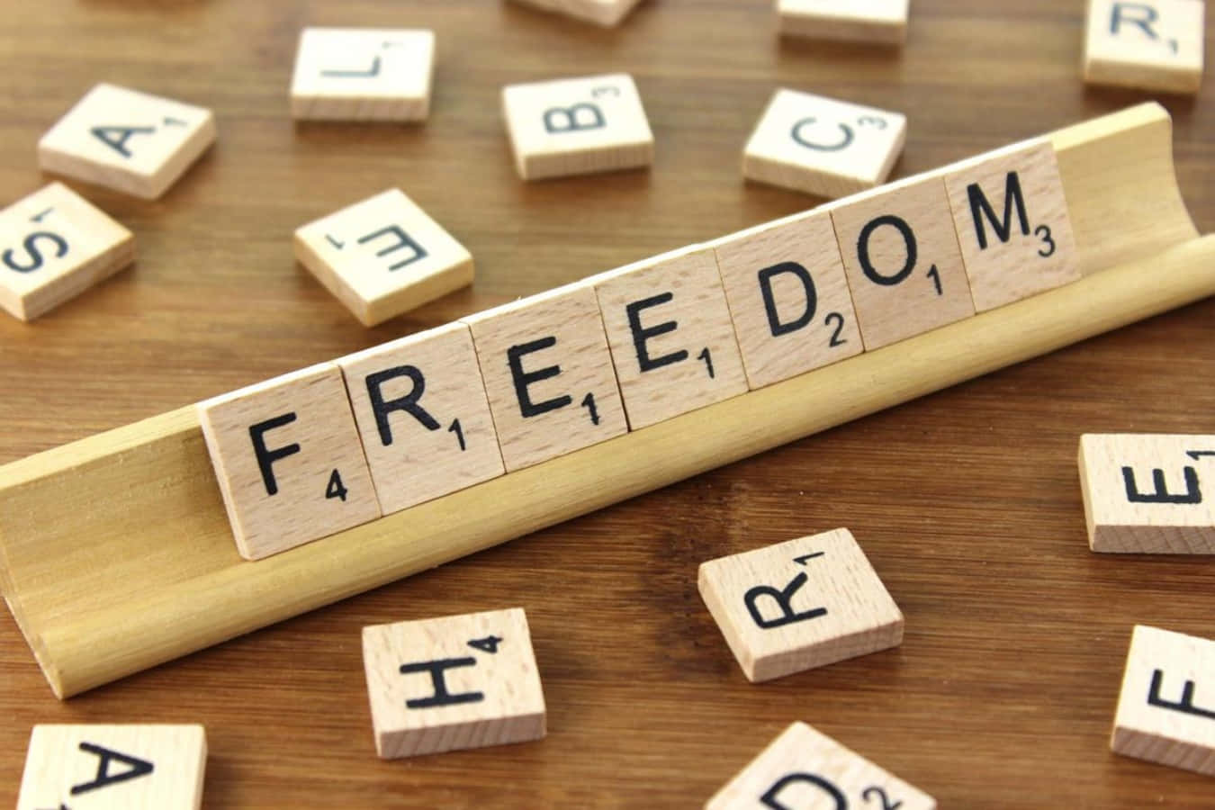 自由 桌布 壁紙 — 木製 Scrabble 字母磚拼出 freedom，放在木架上，置於溫暖的棕色桌面上，周圍散落著字母。