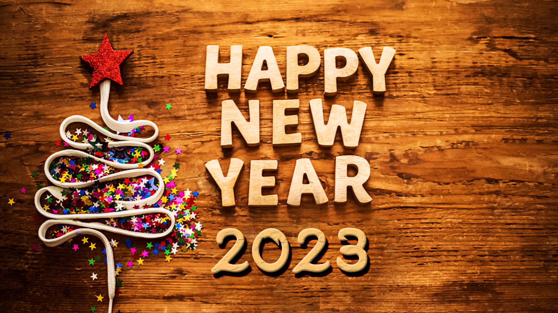 新年 桌布 壁紙 — 木製字母拼出新年快樂2023，旁邊有一個用絲帶捲成的樹，頂部有紅色星星，並在質樸的木頭上散落著閃粉。