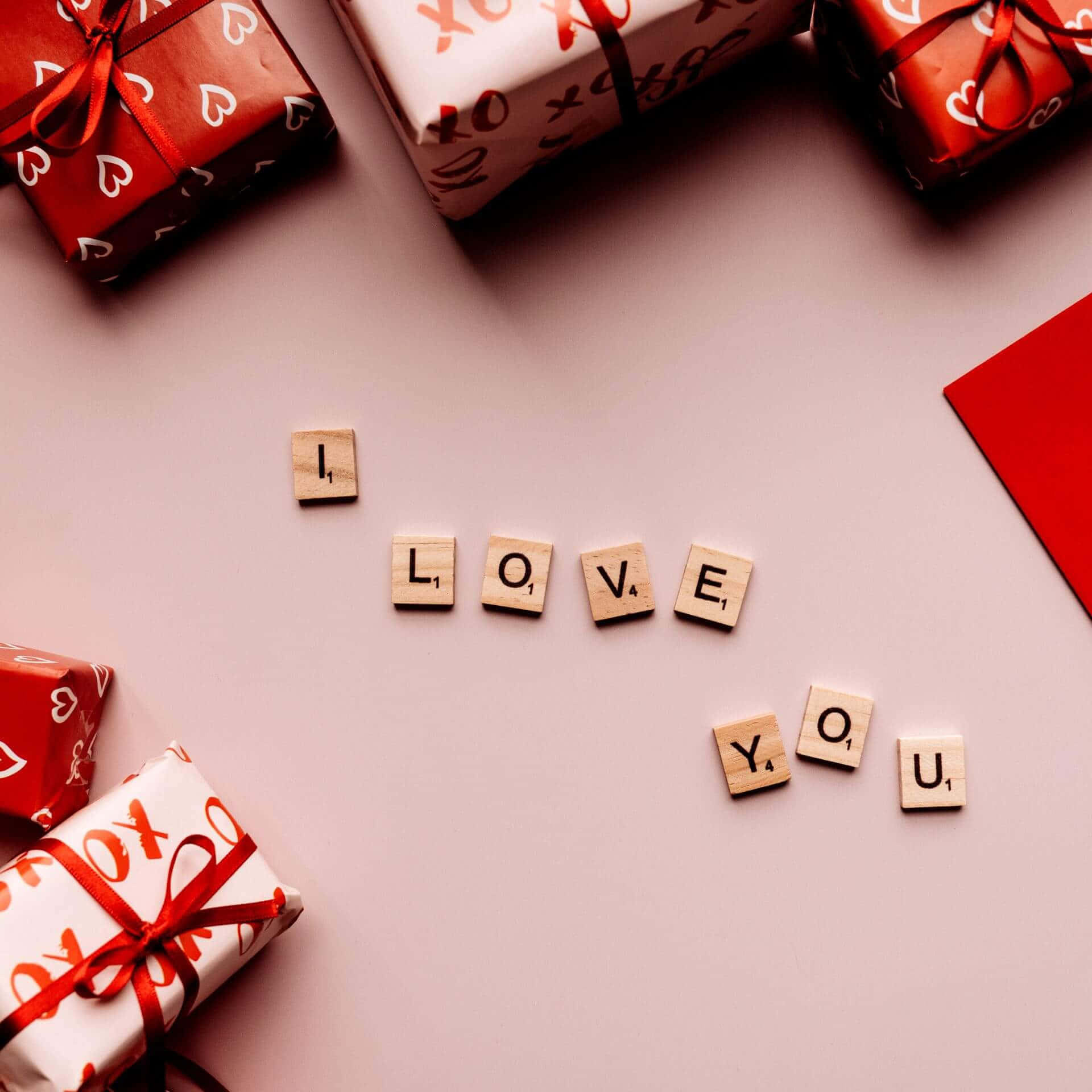 我愛你 桌布 壁紙 — 木製拼字磚拼出「I LOVE YOU」，周圍是紅白心形圖案禮物，放在柔軟粉紅色表面上。