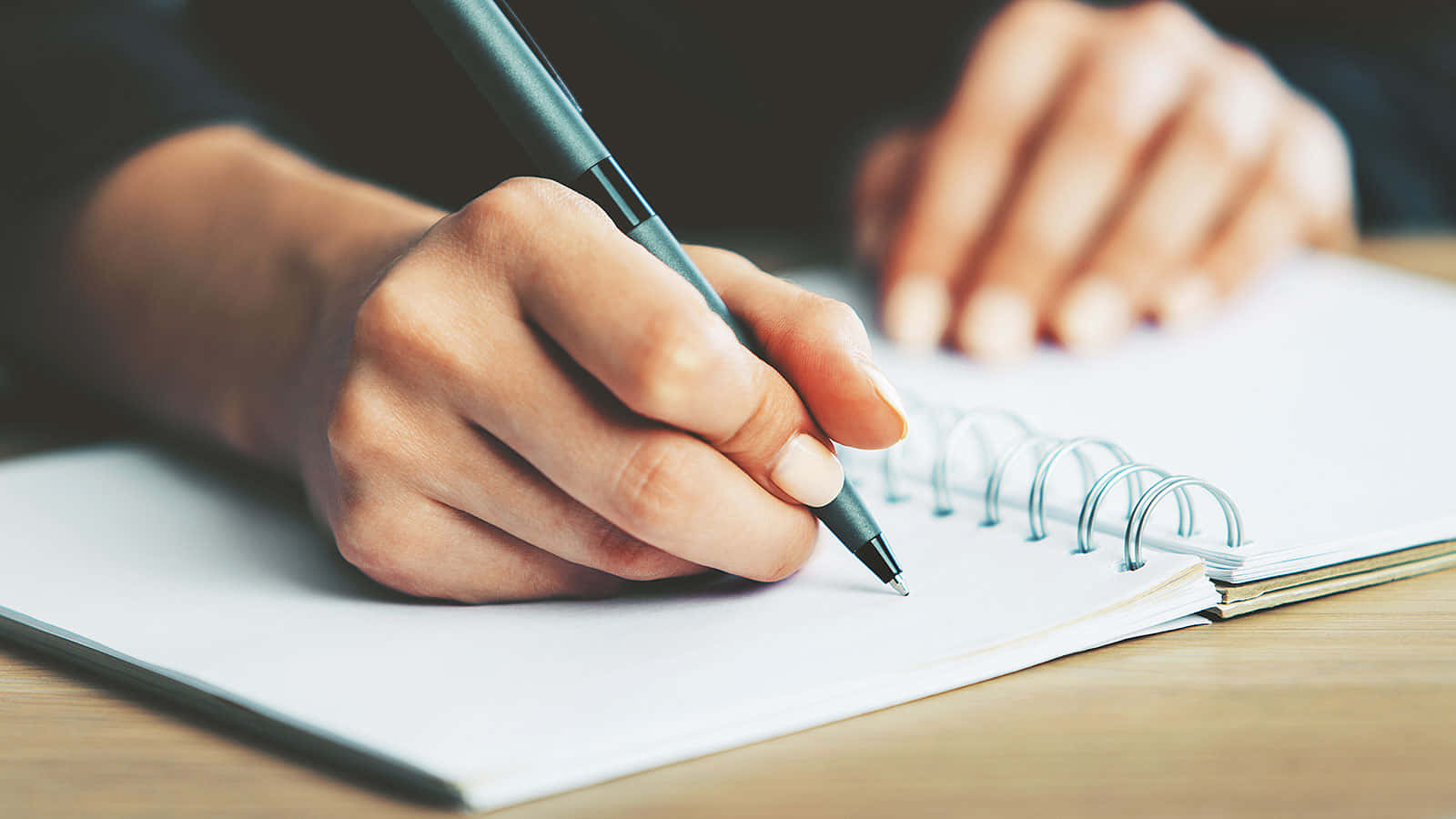 paper 桌布 壁紙 — 木製書桌上，一隻手用筆在螺旋筆記本上書寫的特寫。