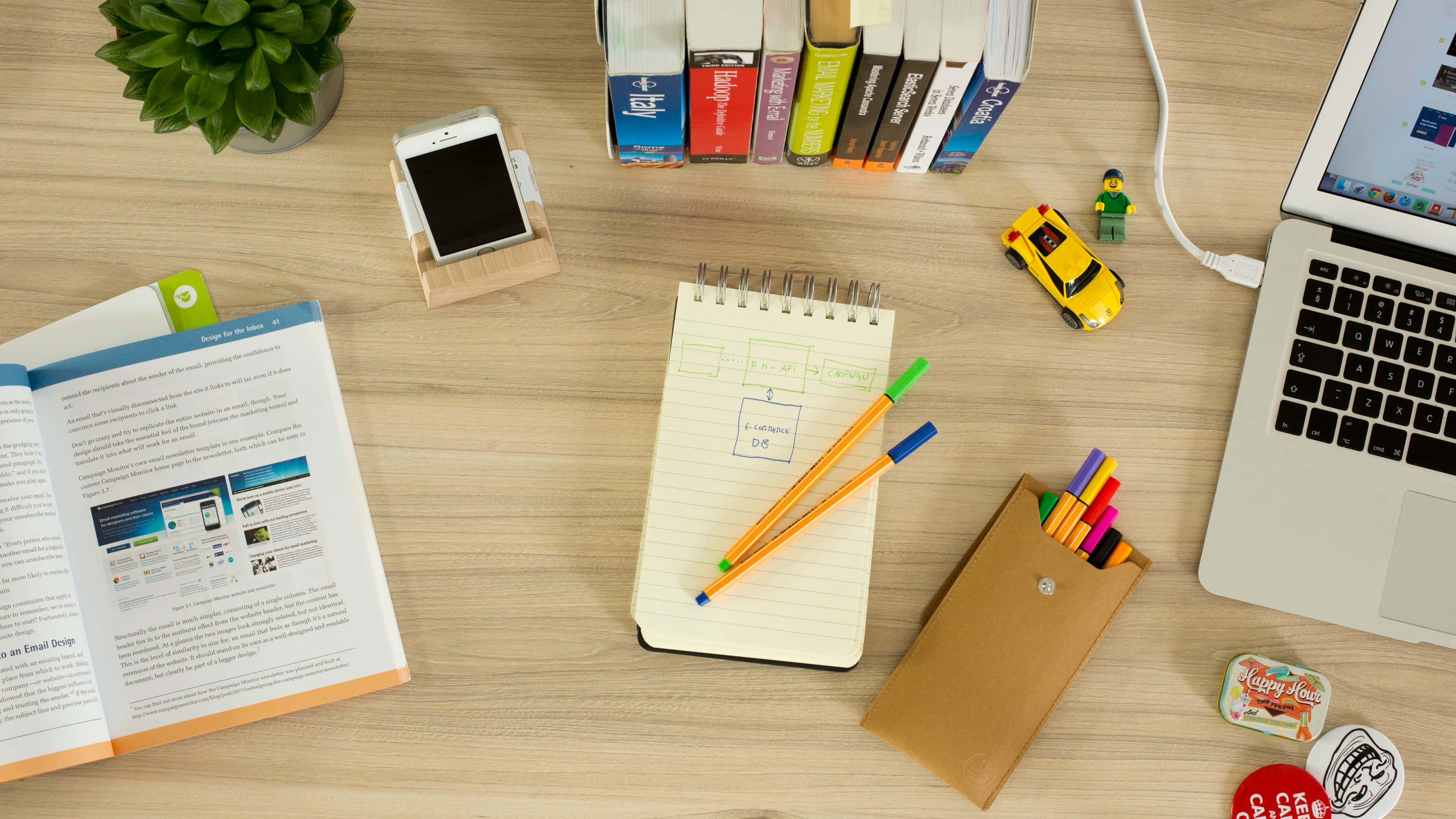 Desk 桌布 壁紙 — 木製書桌的俯視圖，有筆記型電腦、打開的書本、筆記本、鉛筆、彩色筆、電話、書籍和小玩具，營造創意工作空間。