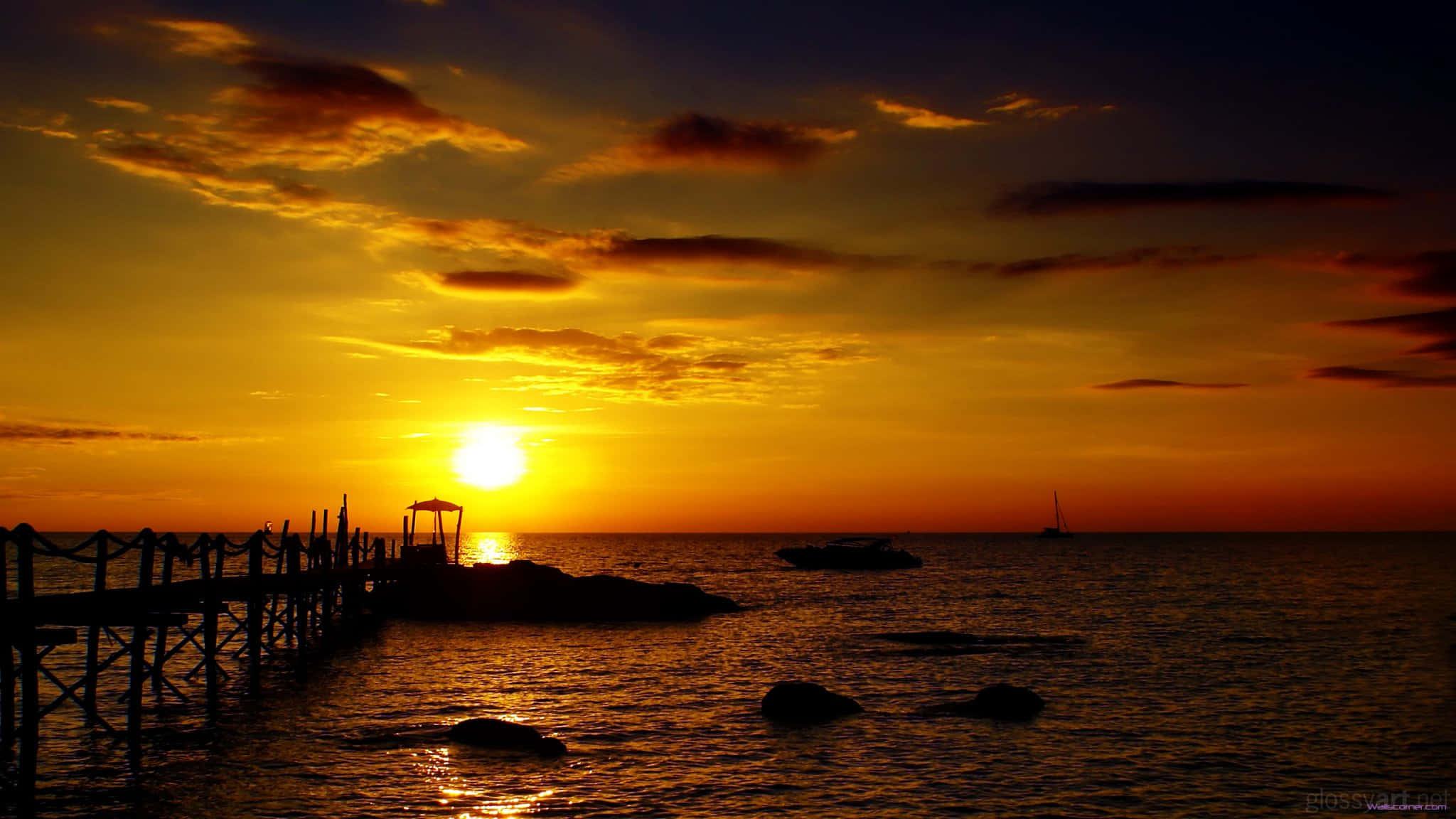 2048x1152 桌布 壁紙 — 木製碼頭延伸至海洋，夕陽以火熱的橙色和金色落下，投射出岩石和船隻的剪影。