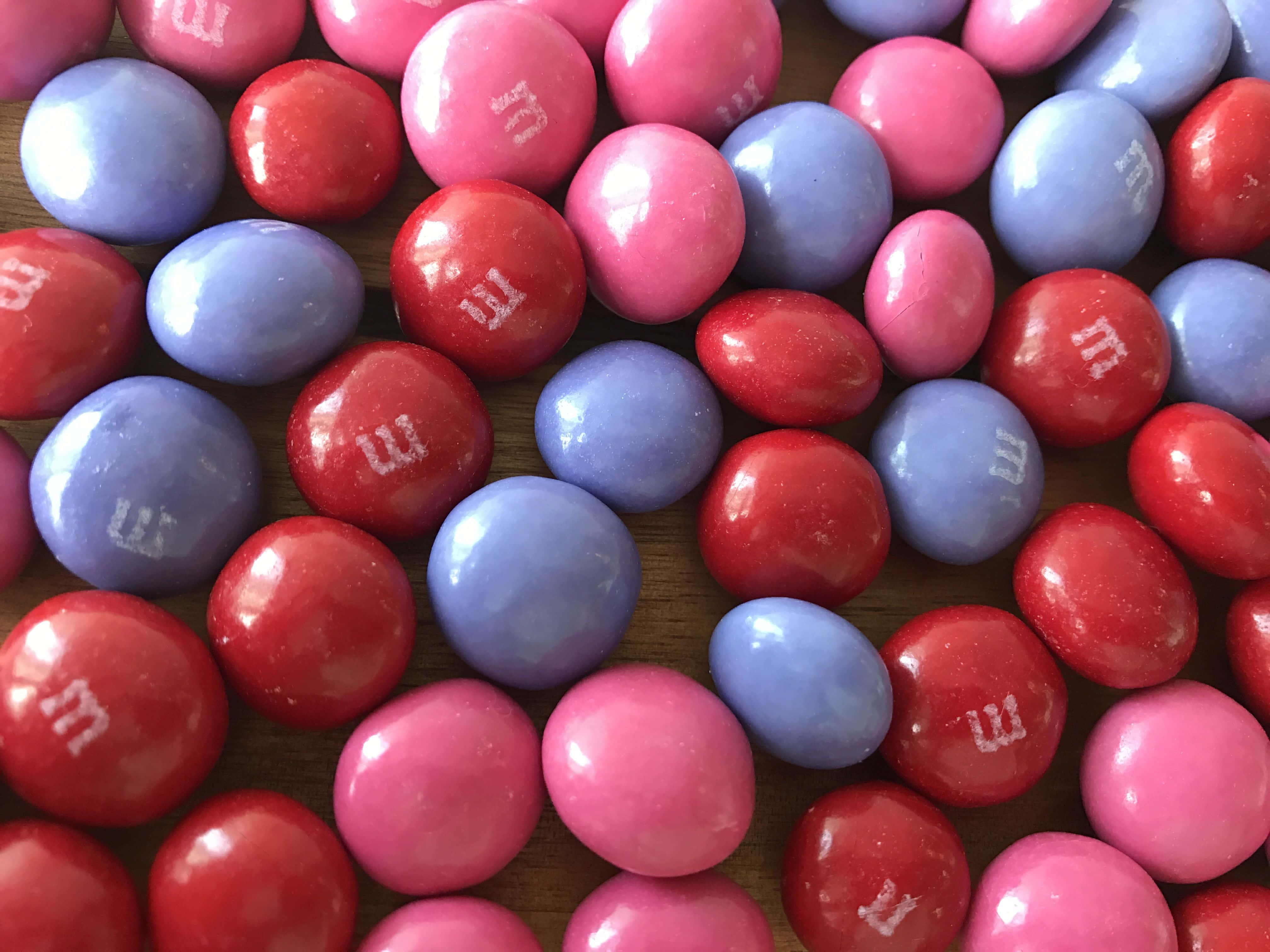 Pink Candy 桌布 壁紙 — 木質表面上散落著光澤的紅色、粉紅色和藍色M&M糖果的特寫。