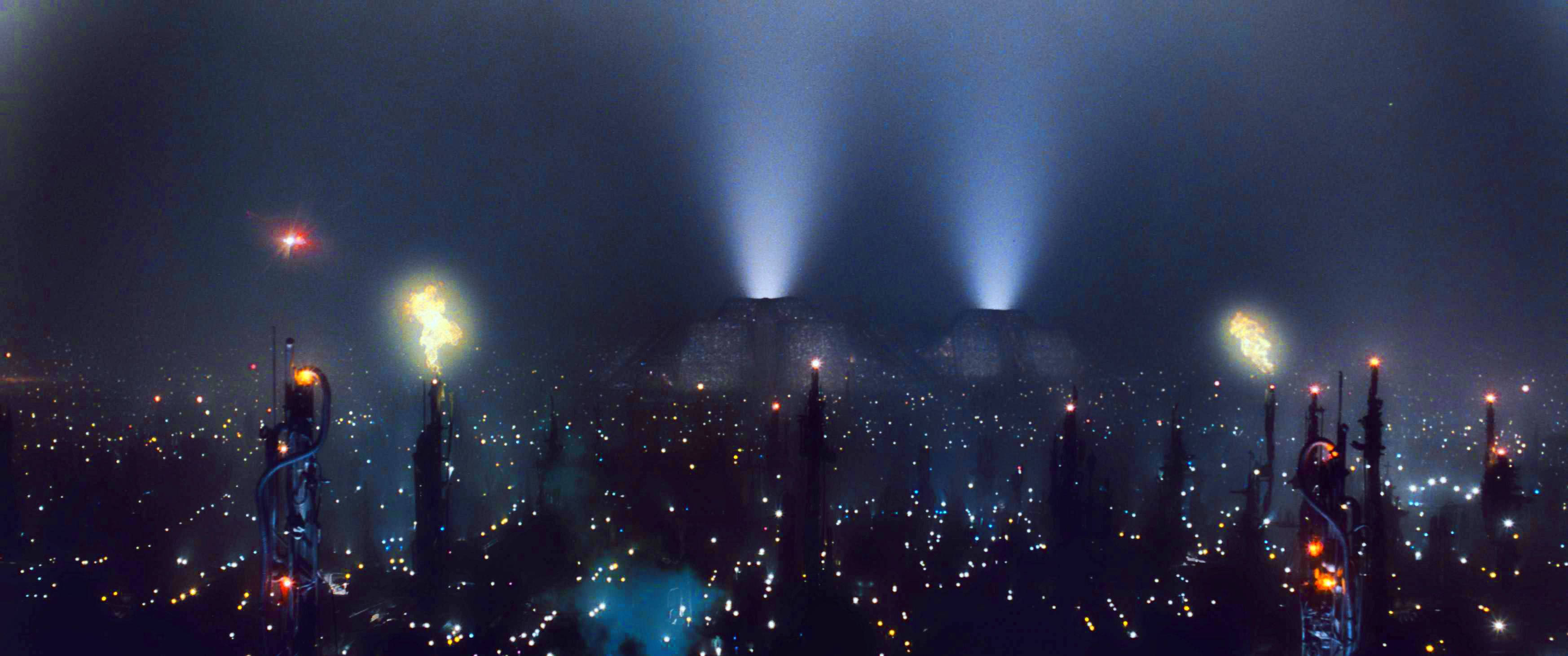 blade 桌布 壁紙 — 未來主義的城市夜景，高聳的尖塔在藍色光束下發光，遠處有熾熱的爆炸。