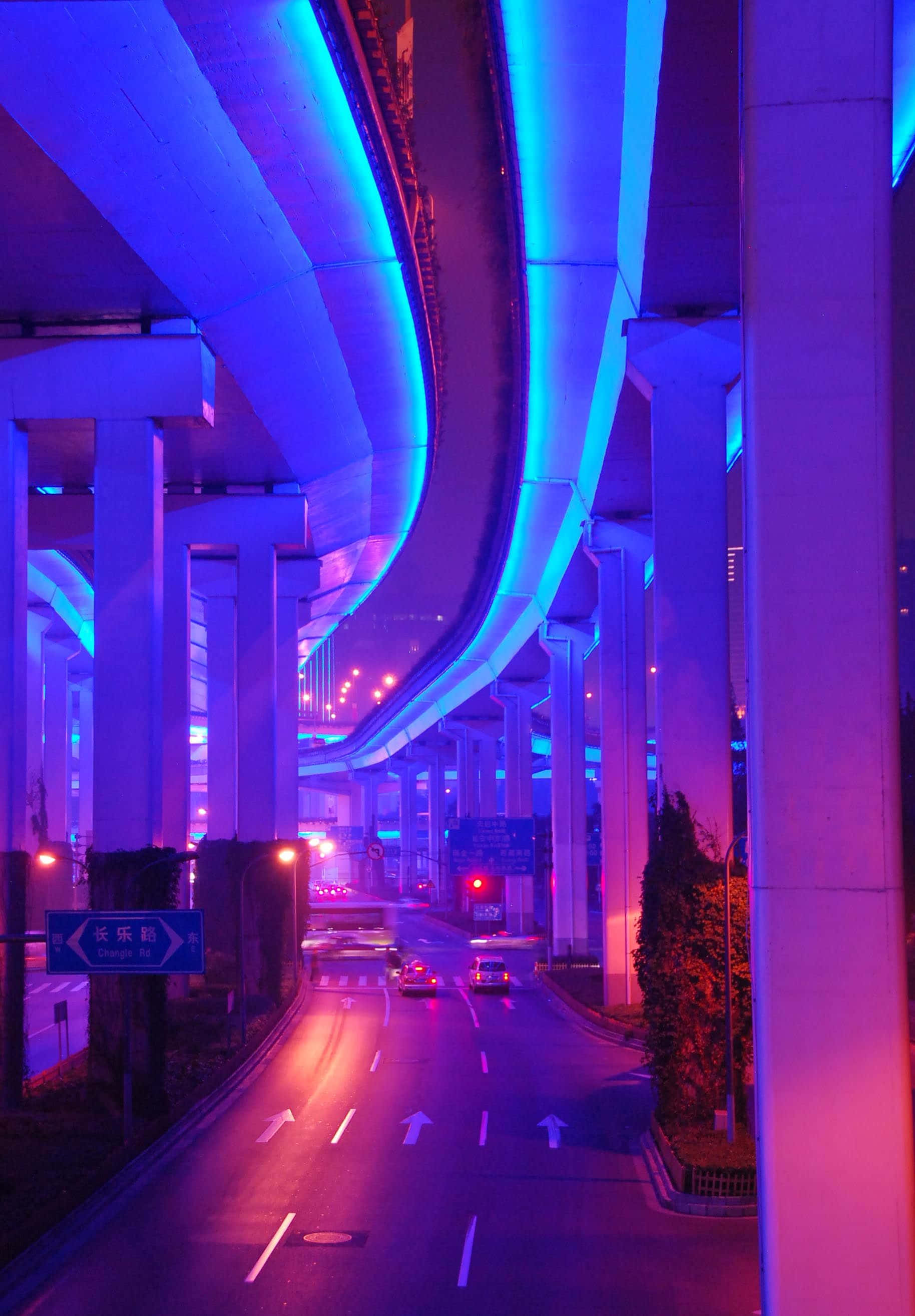 霓虹美學 桌布 壁紙 — 未來主義的城市高速公路夜景，沐浴在鮮豔的藍色和紫色霓虹燈光中，汽車在高聳的混凝土結構下行駛。