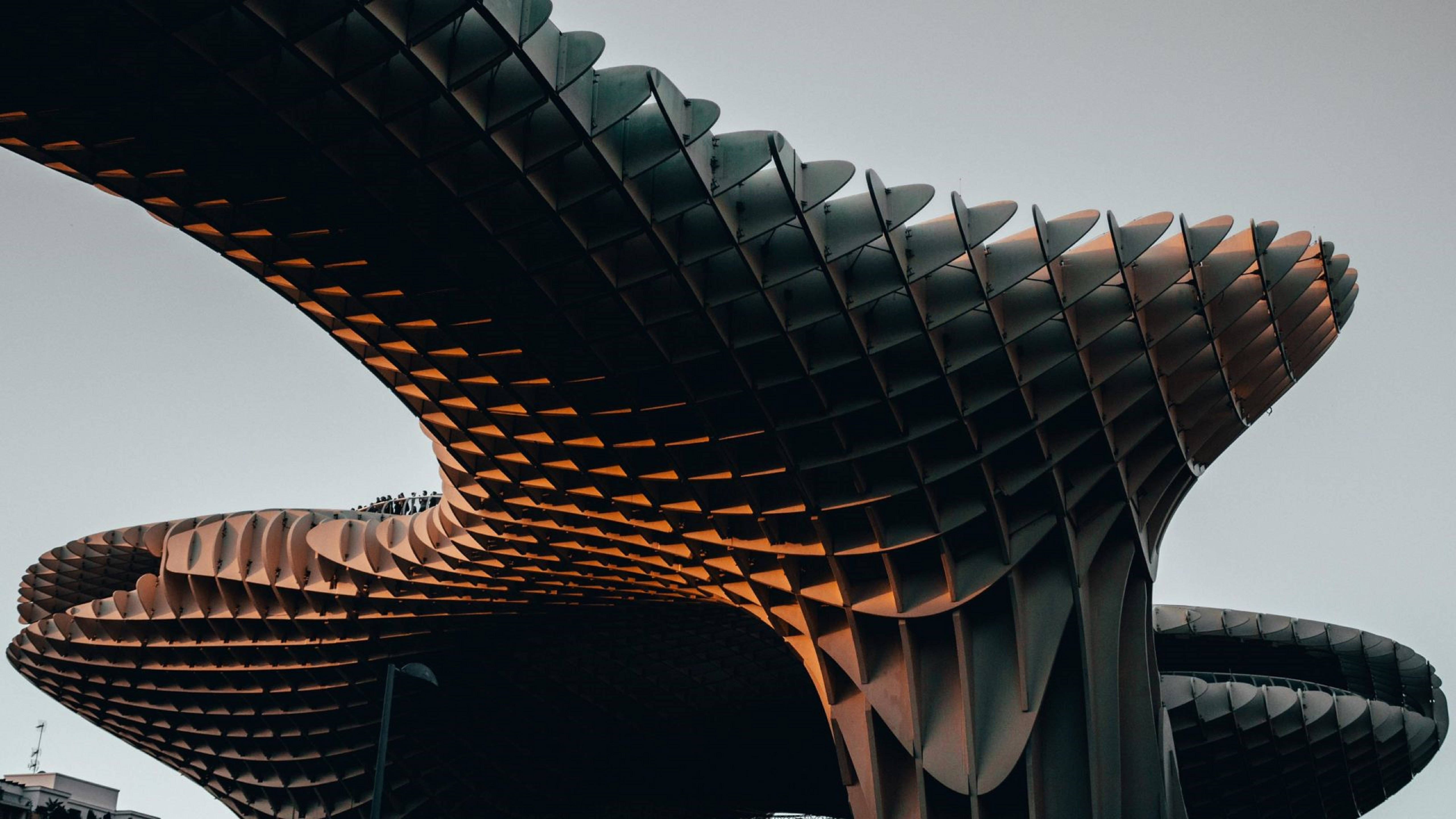 Architecture 桌布 壁紙 — 未來主義的金屬結構，帶有分層的波浪狀面板，在蒼白的天空下溫暖地發光。