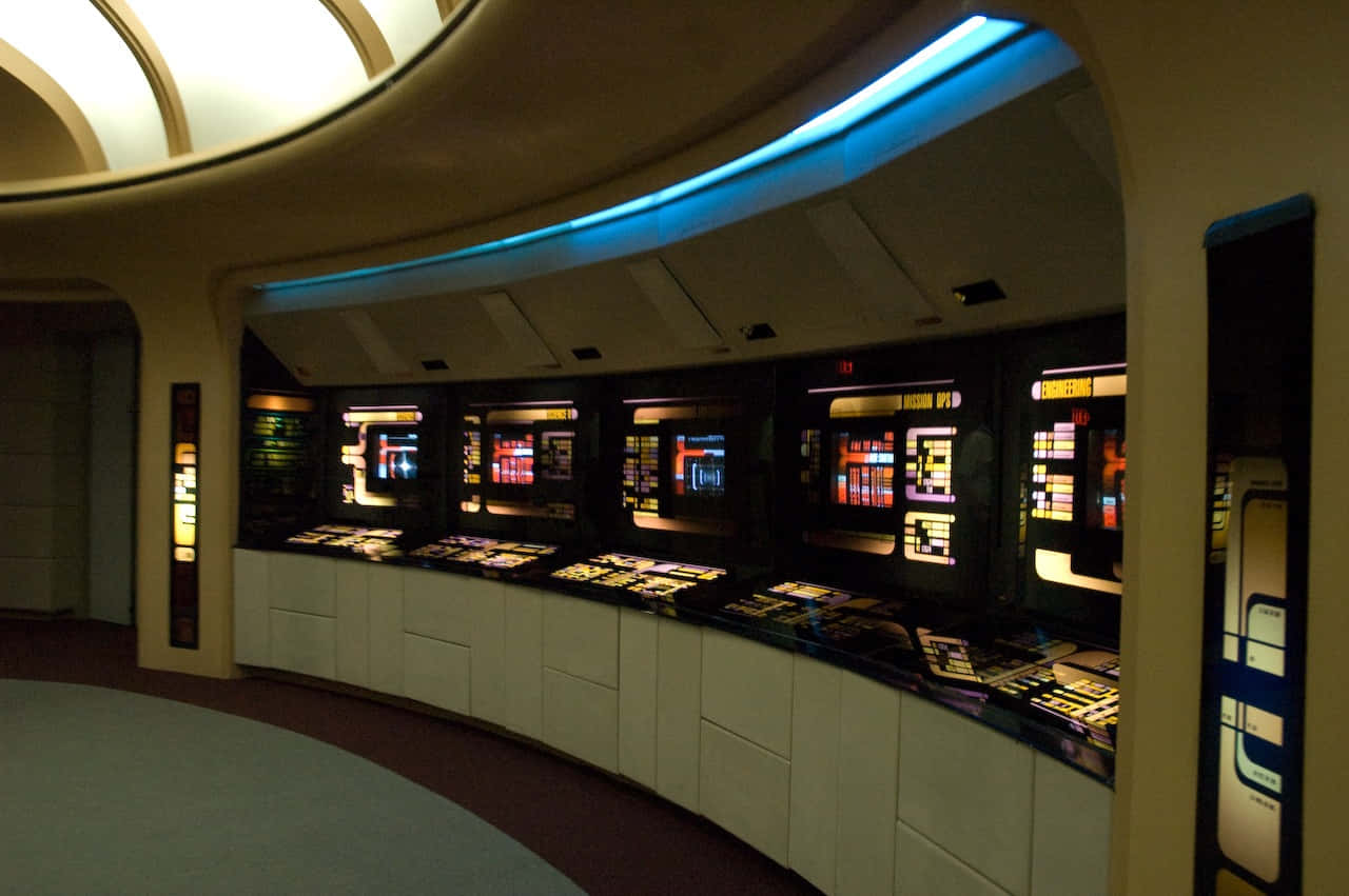 Star Trek Enterprise Bridge 桌布 壁紙 — 未來風格的弧形控制室，帶有發光的藍色霓虹燈和顯示彩色數據介面的照明螢幕。