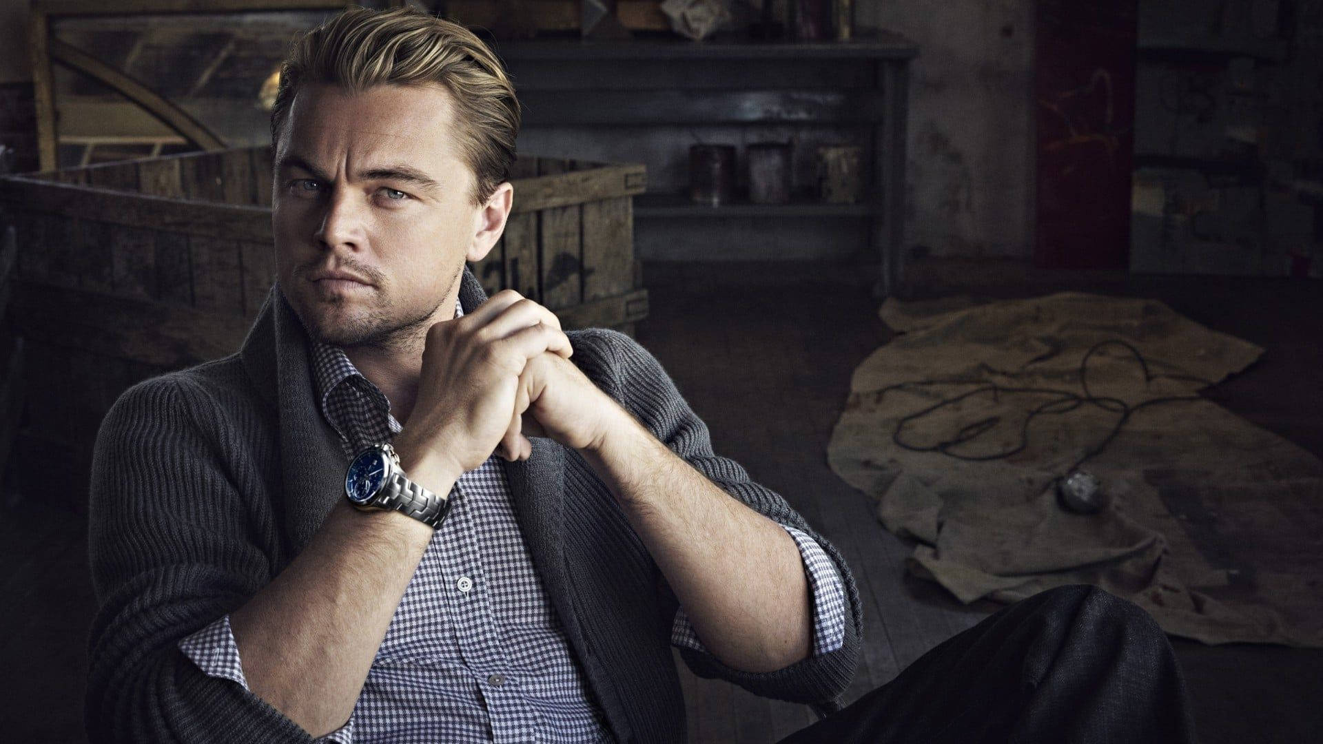Dicaprio 桌布 壁紙 — 李奧納多·狄卡皮歐坐在光線昏暗的鄉村風格房間中，雙手緊握，穿著格紋襯衫和開襟毛衣。